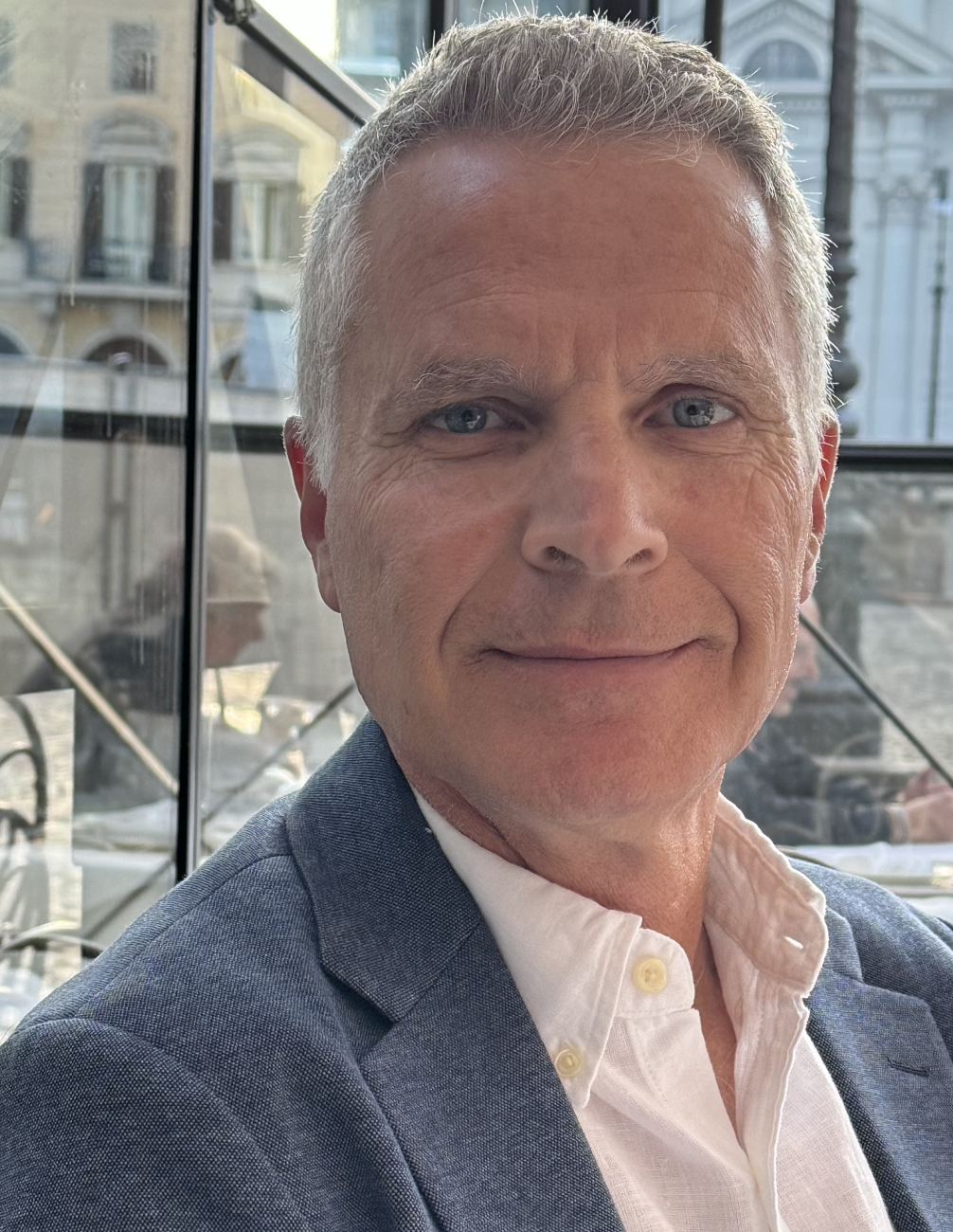 A middle-aged man with gray hair and blue eyes, wearing a light blue suit jacket and a white dress shirt, smiling at the camera. Behind him are city buildings and large windows.