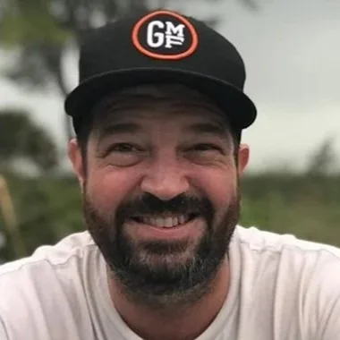 A smiling man wearing a black baseball cap with a red and white logo, outdoors with trees in the background.