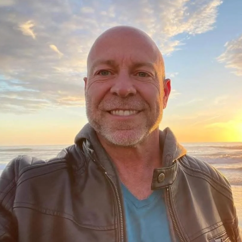 A smiling middle-aged man with a beard taking a selfie on the beach at sunset, wearing a leather jacket and a blue shirt, with the ocean and colorful sky in the background.
