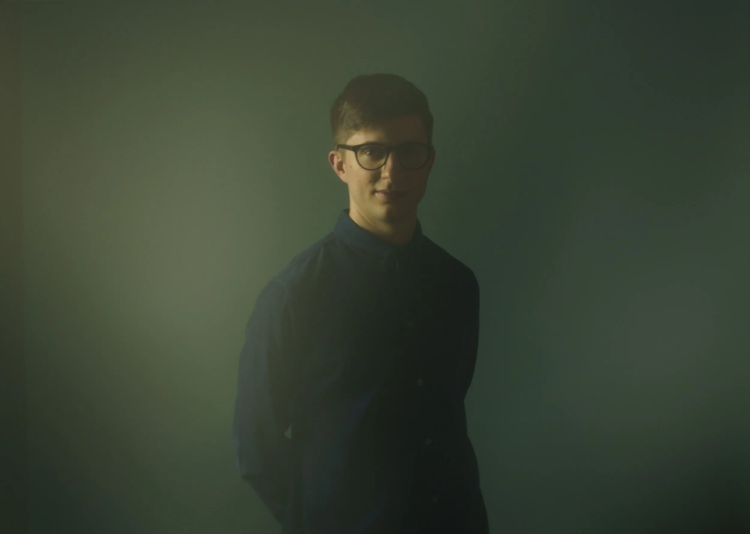 Portrait of pianist and composer Liam Kaplan wearing a dark blue shirt and glasses against a dark green background