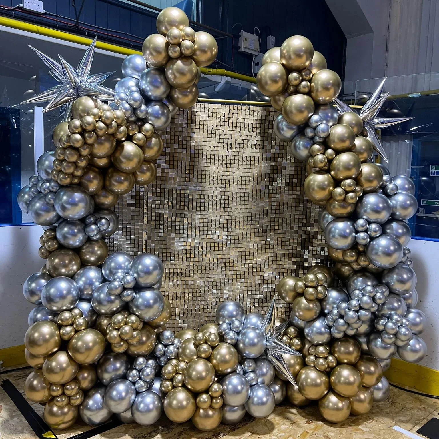 Decorative holiday balloon arch with silver and gold balloons, accented with large silver star-shaped balloons, in front of a gold mosaic backdrop.