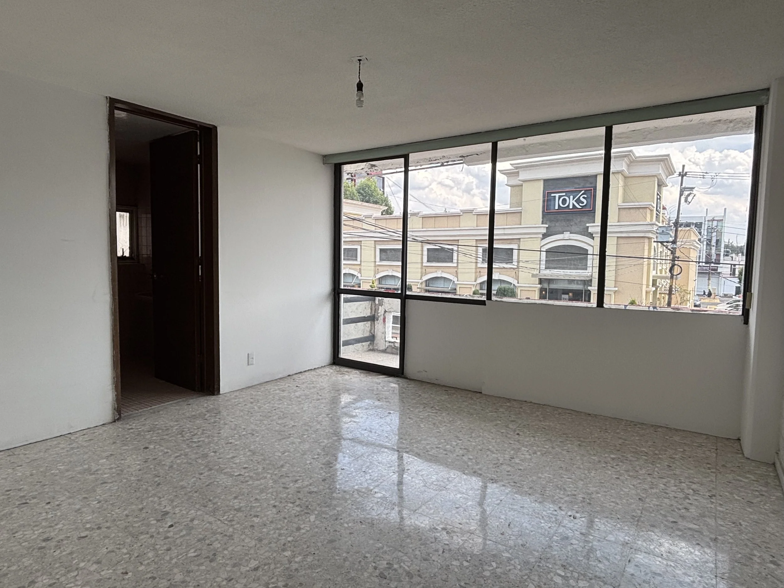 An empty room with terrazzo flooring, beige walls, a large window with a sliding door, and a view of a commercial building labeled "Toks" outside. There is an open doorway on the left with a dark room beyond. The ceiling has a single light bulb hangi