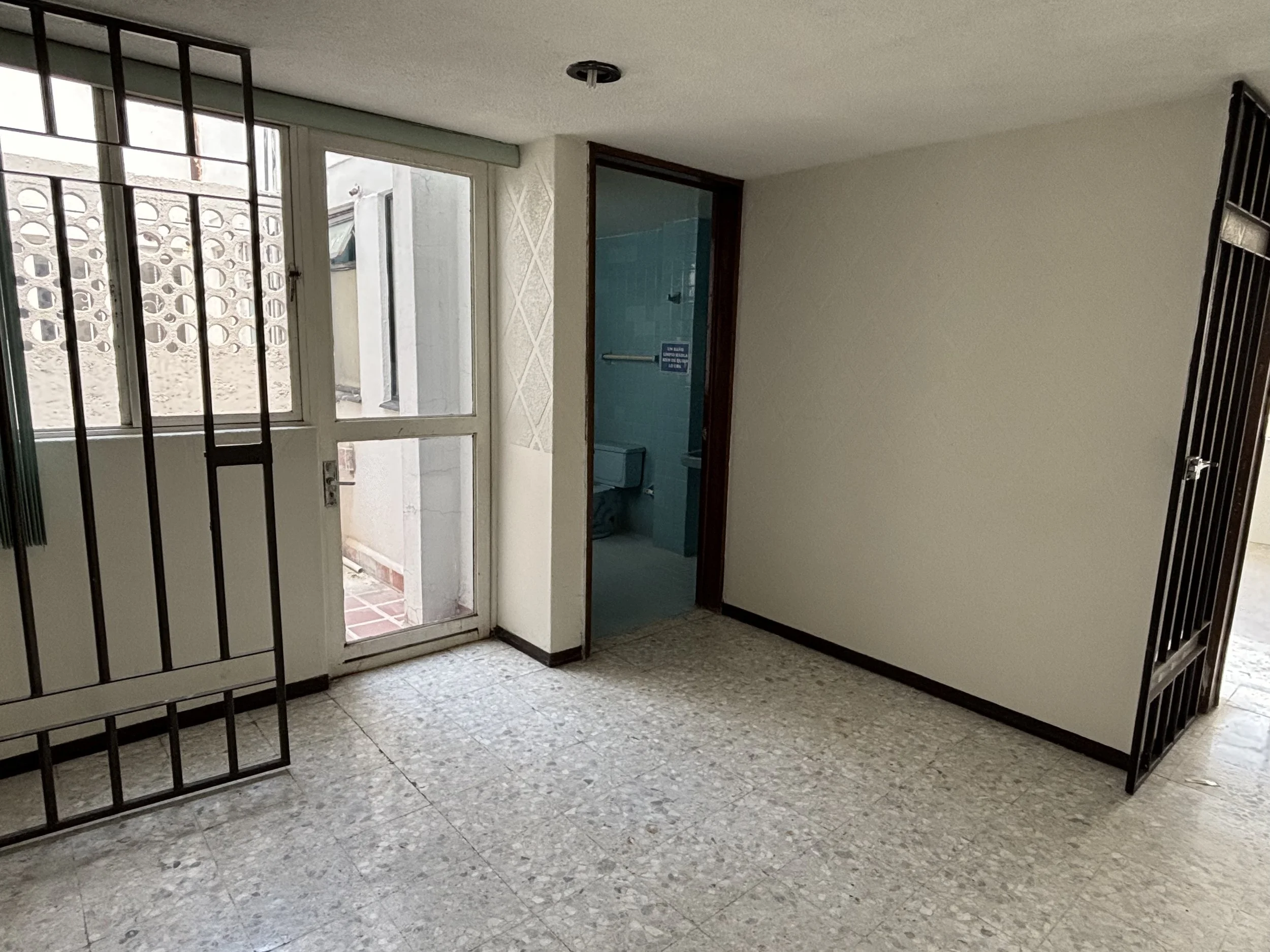 An empty room with a window and door leading outside, a small bathroom with a toilet, and metal bars on the windows and door.