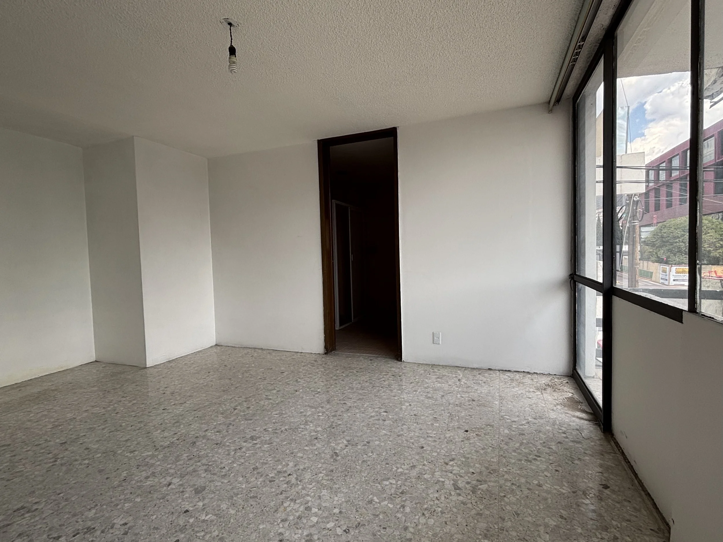 Interior of an empty room with white walls, a terrazzo floor, a windowed wall on the right, and a doorway on the back wall.