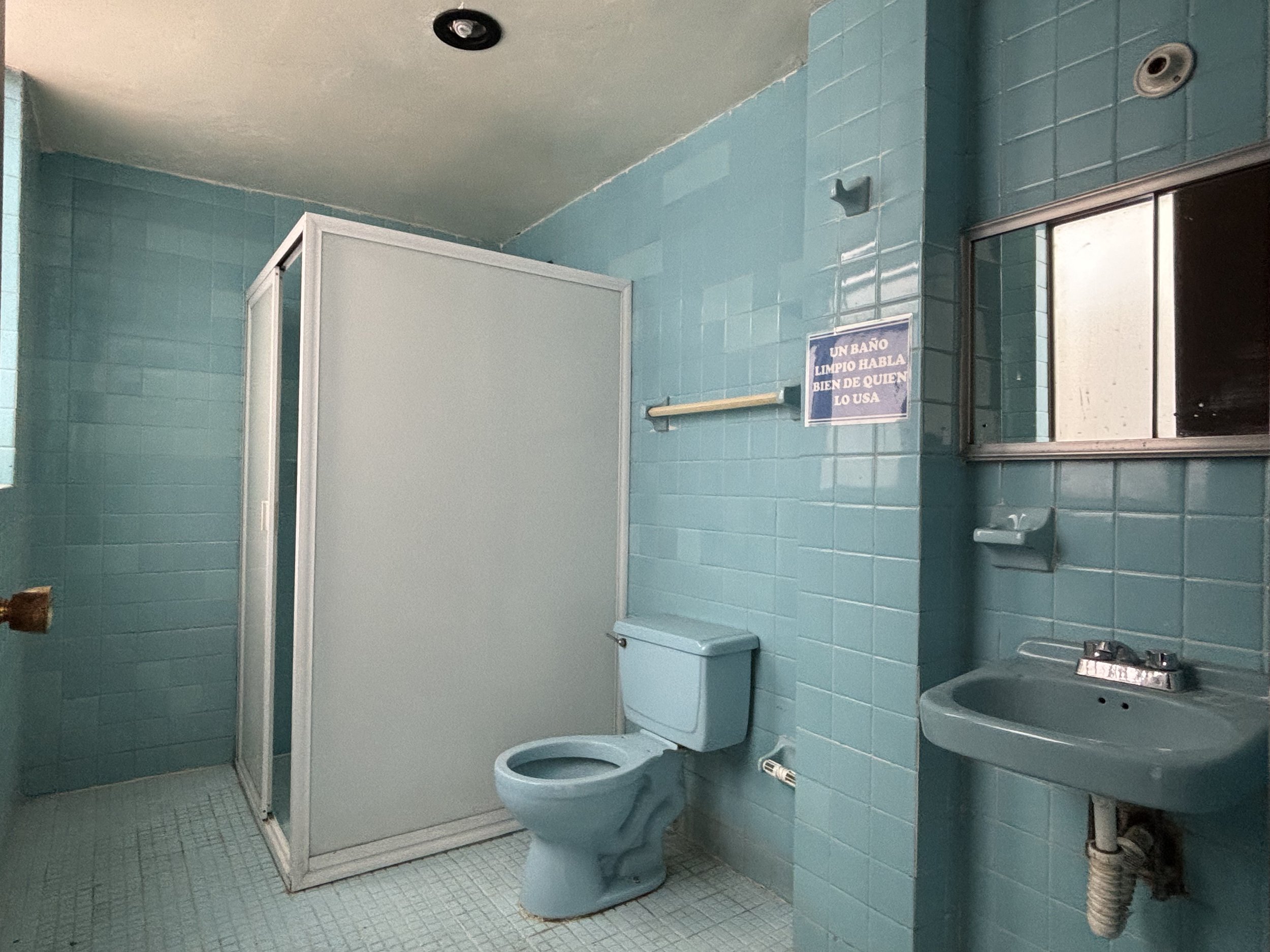 A small bathroom with light blue tiles on the walls. It has a toilet, a small matching sink, a mirror, and a shower stall with a sliding door. There is a sign on the wall with text in Spanish.