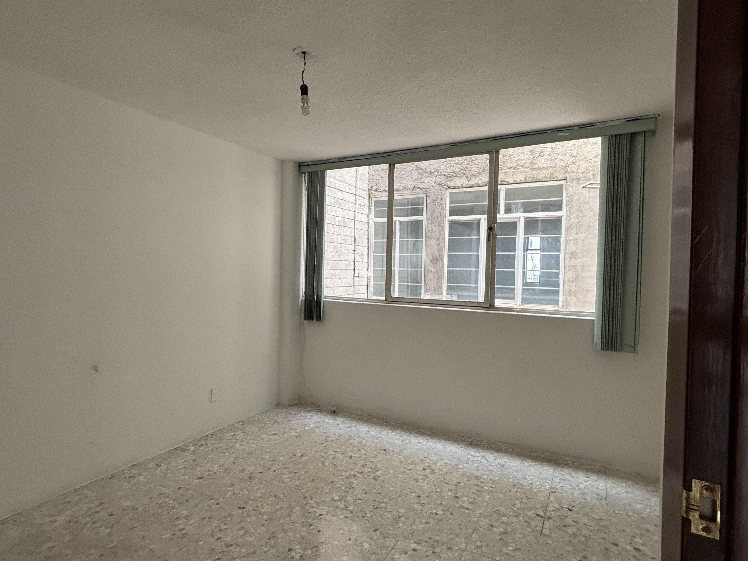 Empty room with white walls, a large window with vertical blinds, a light bulb hanging from the ceiling, and a speckled tile floor.