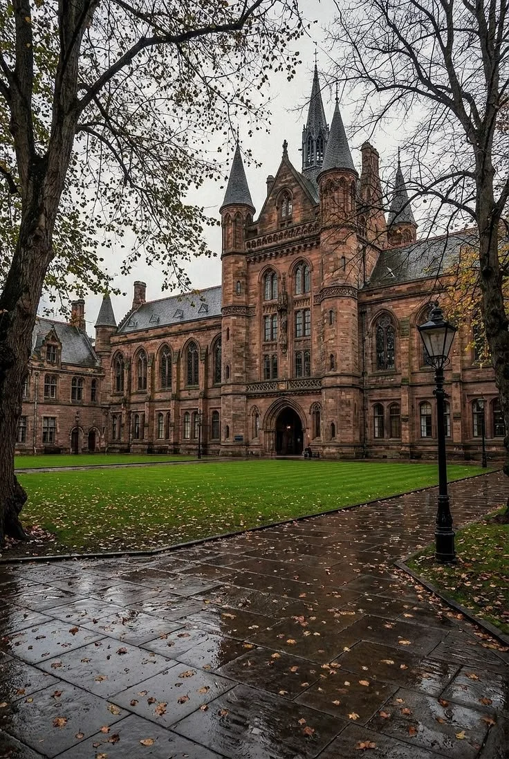 A historic castle-like building made of red sandstone with pointed towers, surrounded by leafless trees and a wet pathway with fallen leaves.