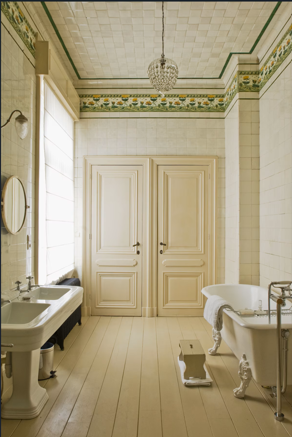 A vintage-style bathroom with a clawfoot bathtub, dual sinks, a mirror, and a chandelier, with a decorative border on the upper walls and closed paneled doors.