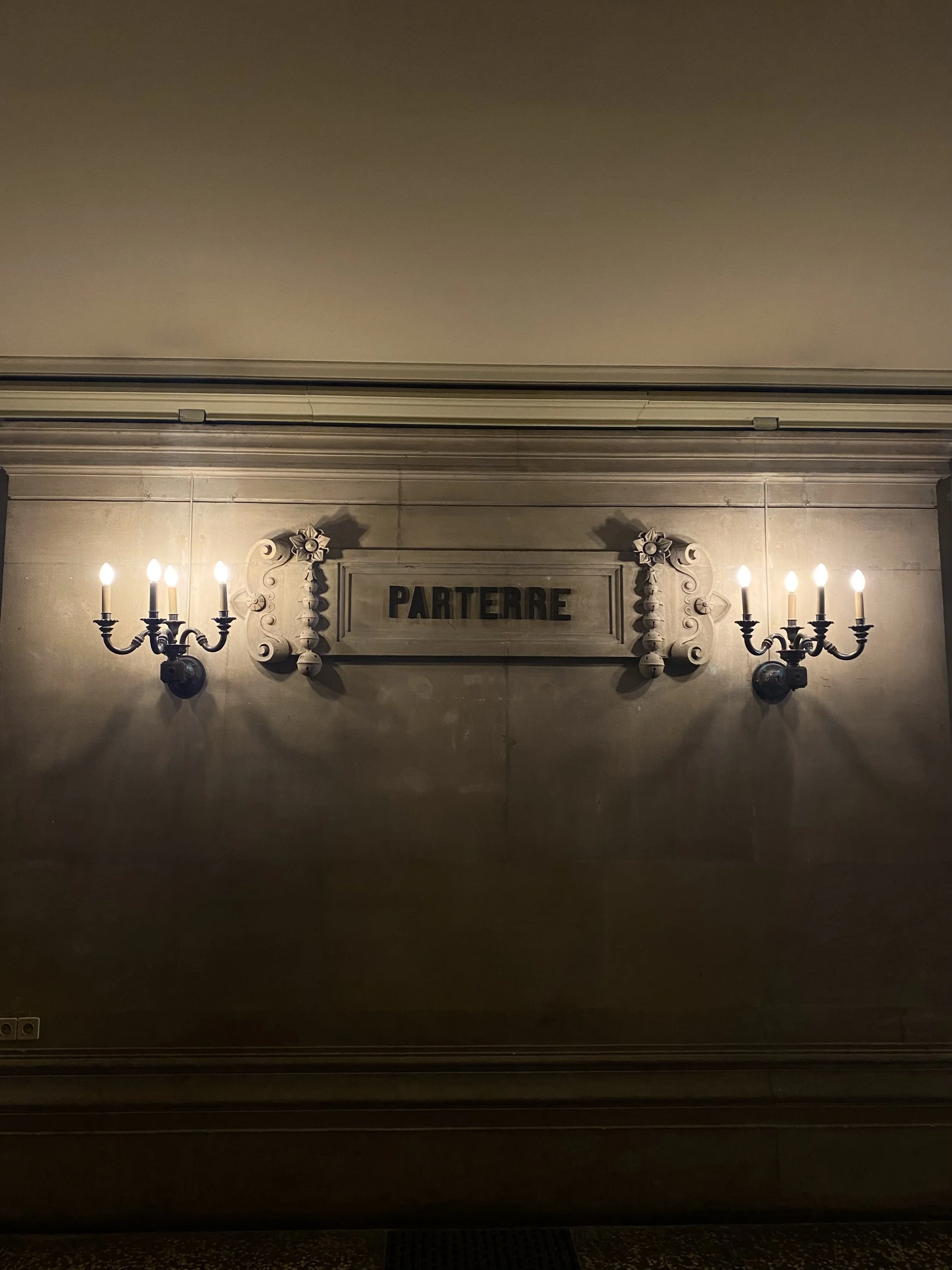 Interior of a building with a beige wall, a framed sign that says 'Parterre', and two ornate wall-mounted candelabra-style light fixtures with five lit candles each.