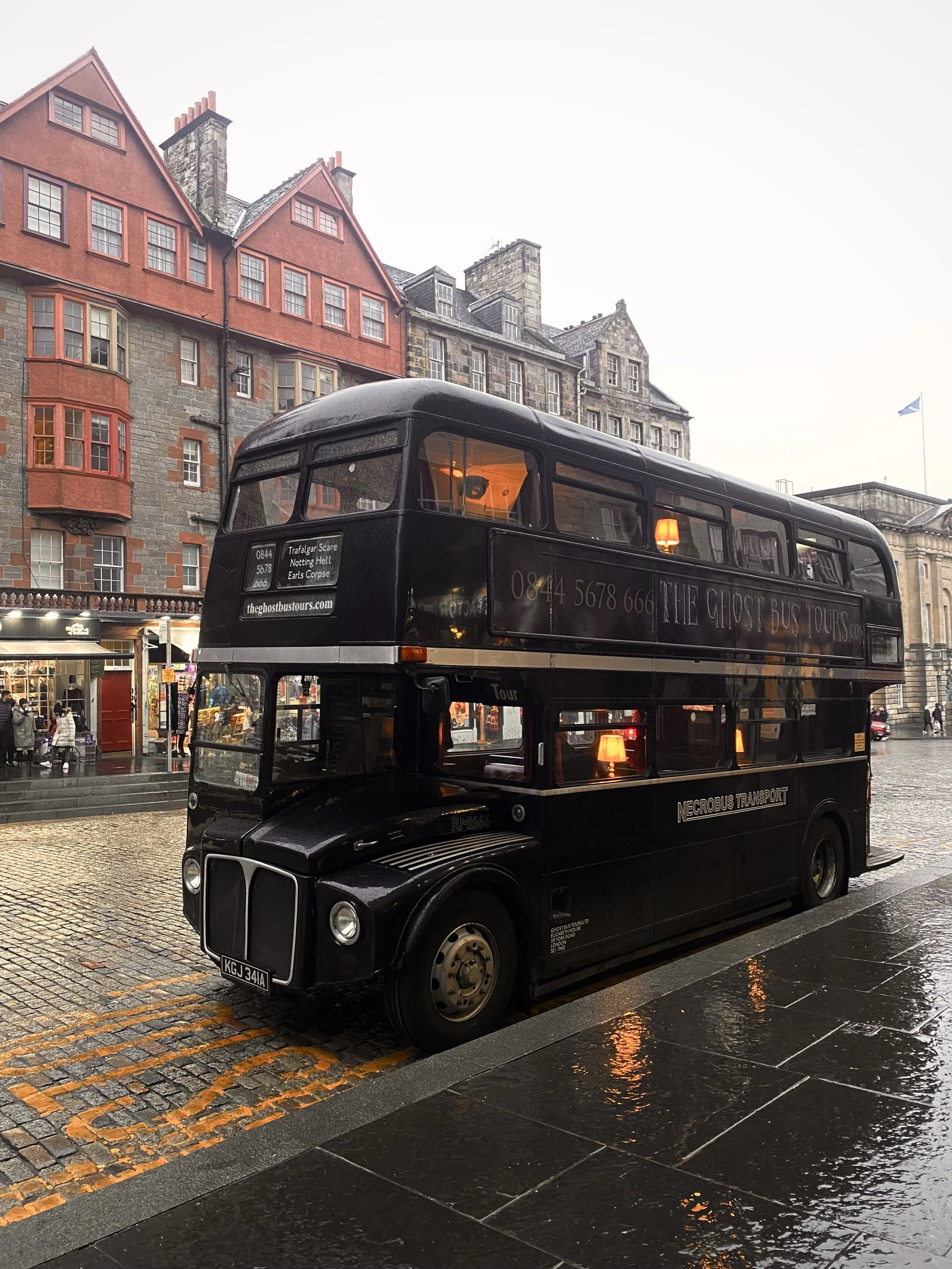 A double-decker black ghost tour bus parked on a wet cobblestone street in front of multi-story historic buildings with red and gray stone facades in a city in the rain.