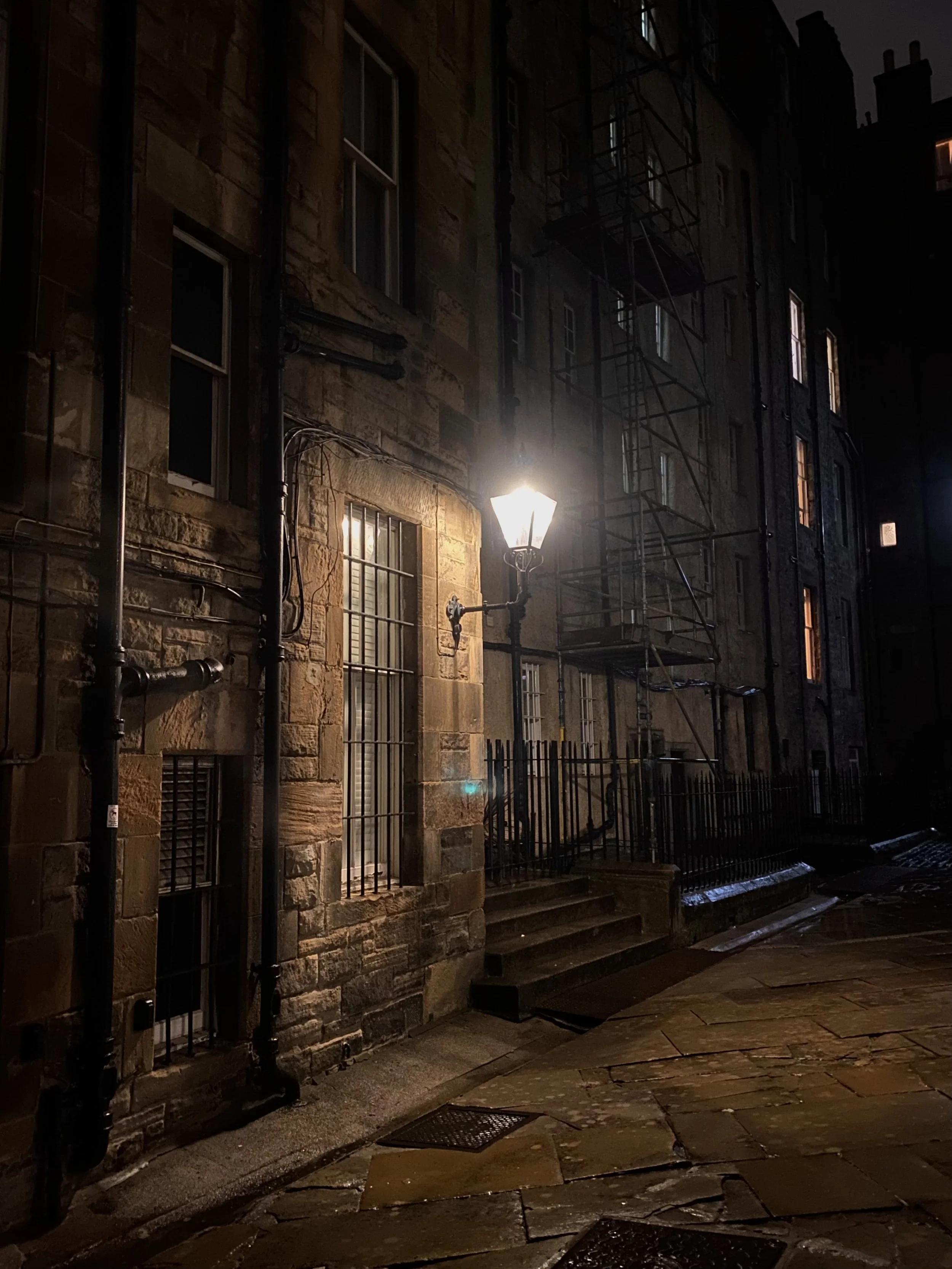 Night scene of a stone building with illuminated windows and a streetlamp, some windows have bars, and there are fire escape scaffolds on the building's exterior.