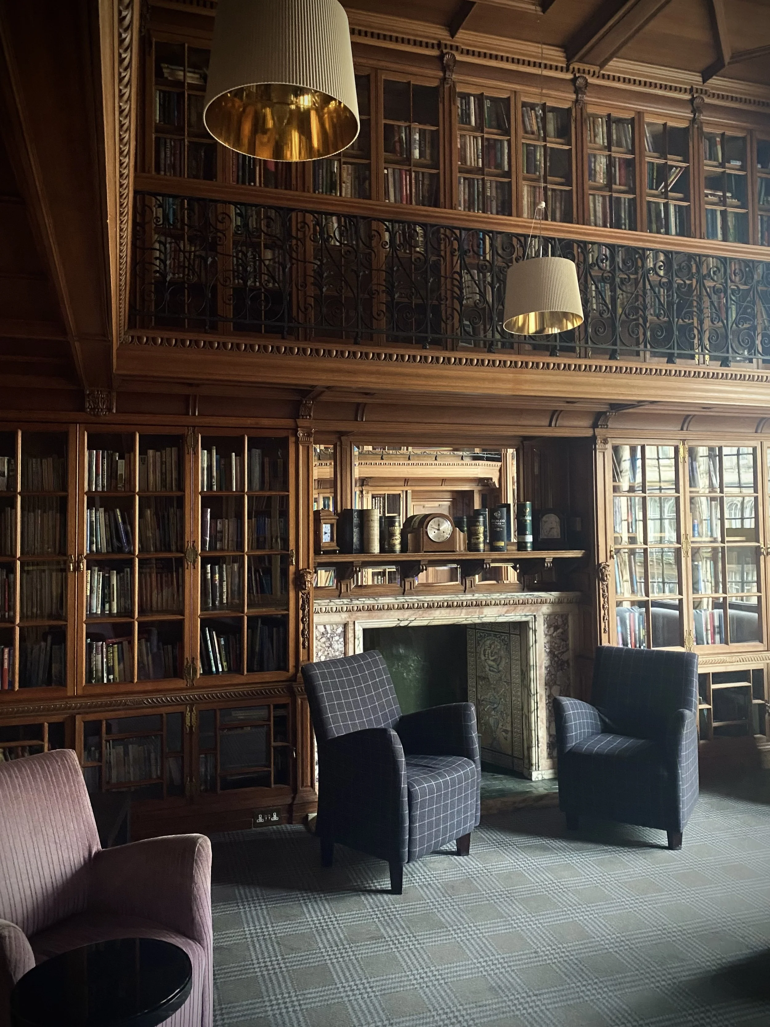 A cozy, elegant library room with wooden bookshelves filled with books, a fireplace, and comfortable chairs with patterned fabric. There are two hanging lamps with gold interiors and a patterned carpet on the floor.