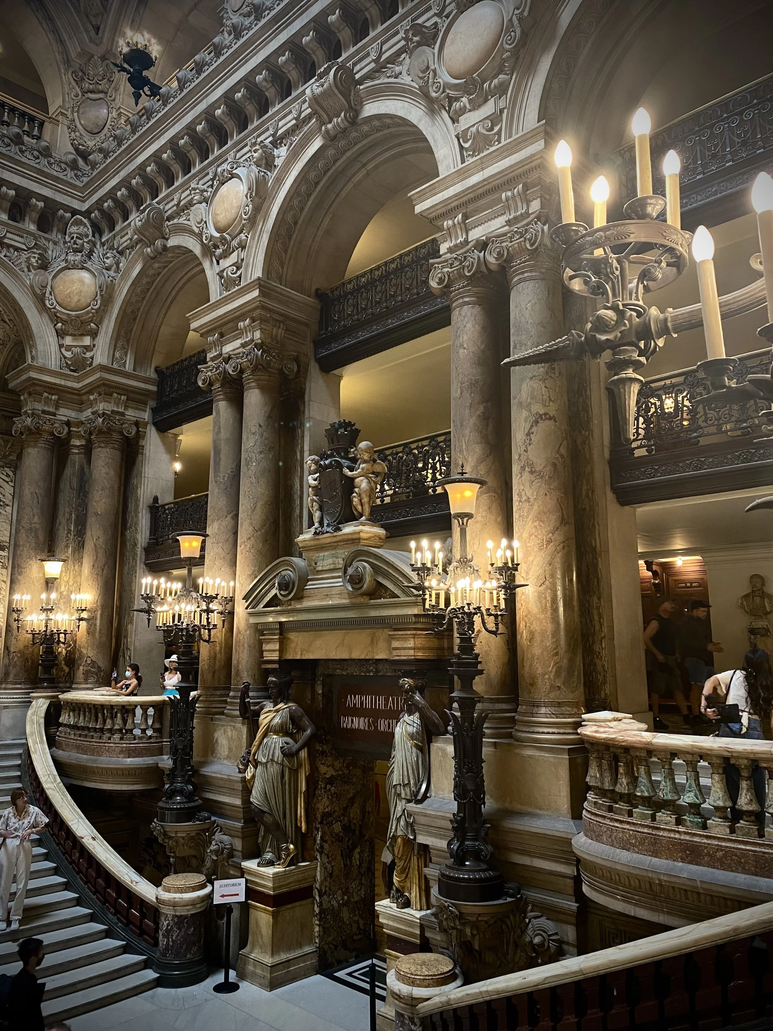 Interior of a grand historic building with large marble columns, ornate architecture, sculptures of women, and multiple chandeliers with lit candles. People are walking and sitting on benches.