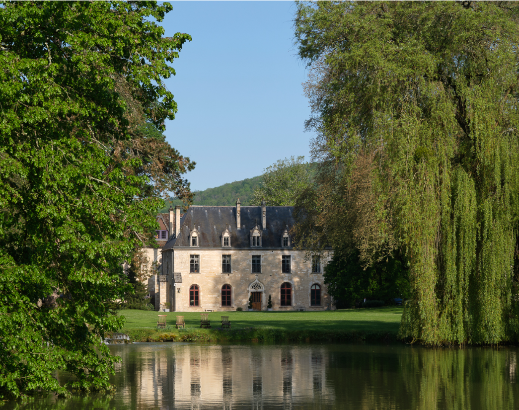 A historic stone mansion surrounded by lush green trees and grass, with a calm pond reflecting the house and trees in the foreground.
