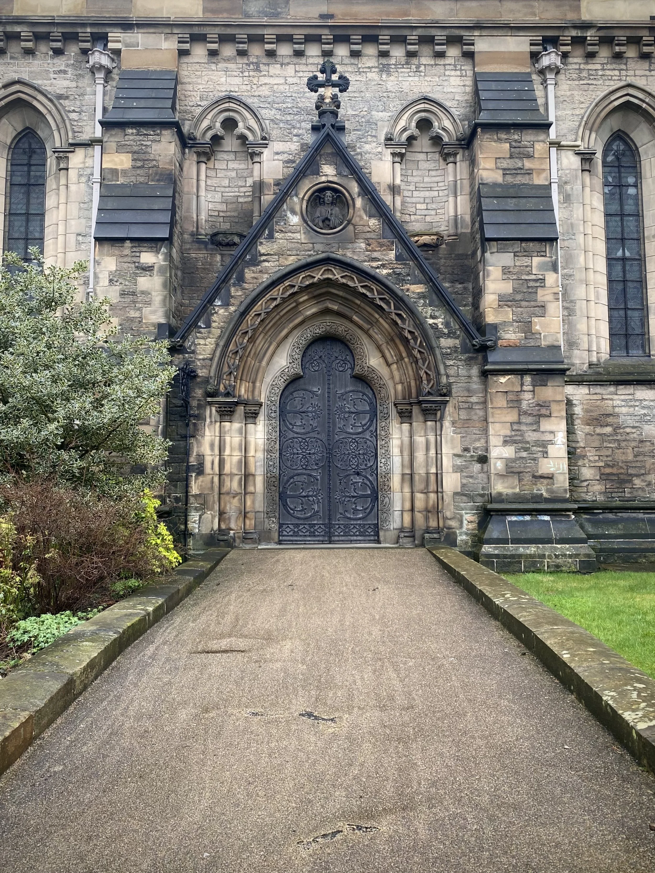 The entrance to a historic stone church with ornate black iron double doors, decorative stone framing, and a carved circular medallion above the doorway. The church has tall, narrow arched windows with stained glass, and a large cross on top of the t