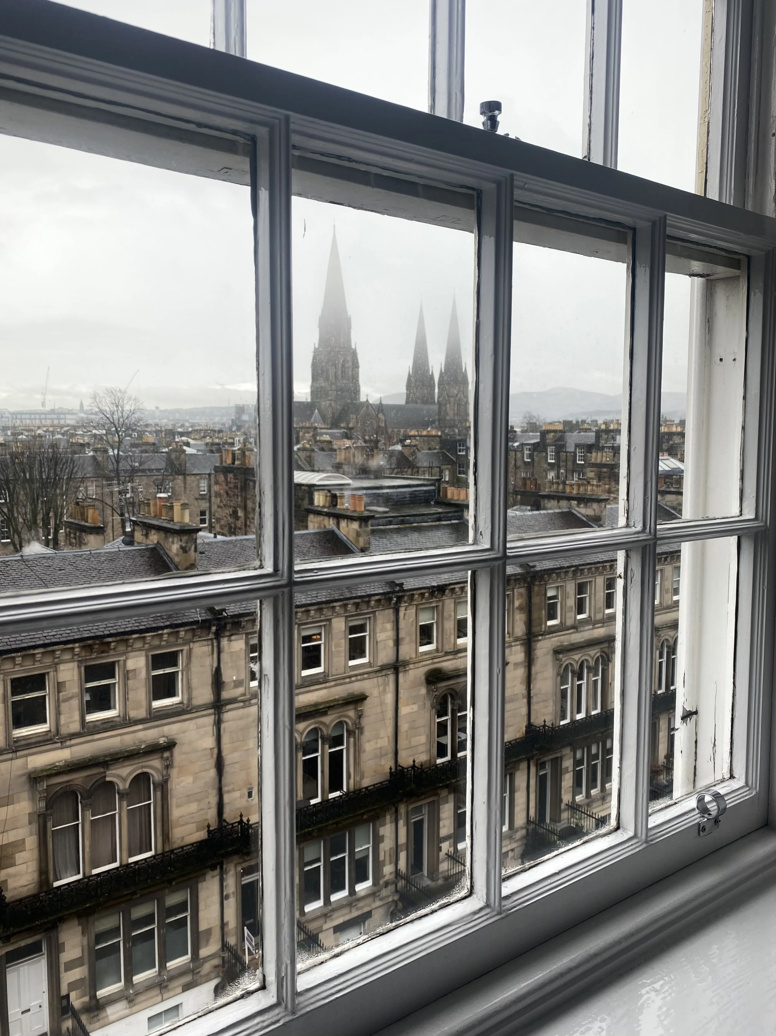 View of city rooftops, with a large church and three prominent church spires seen through a window with white moldings.