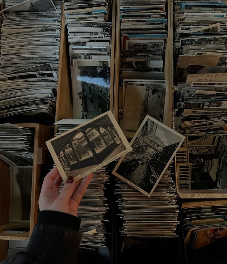 Person holding black and white photographs of cityscapes or streets, with numerous other photographs organized in wooden containers in the background.