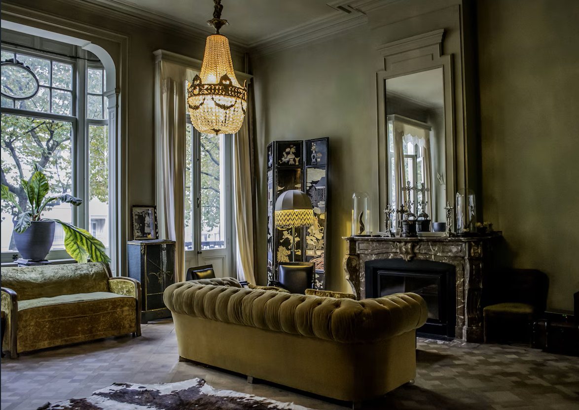 Elegant living room with vintage furniture, a chandelier, large windows, and decorative items on a fireplace mantel.