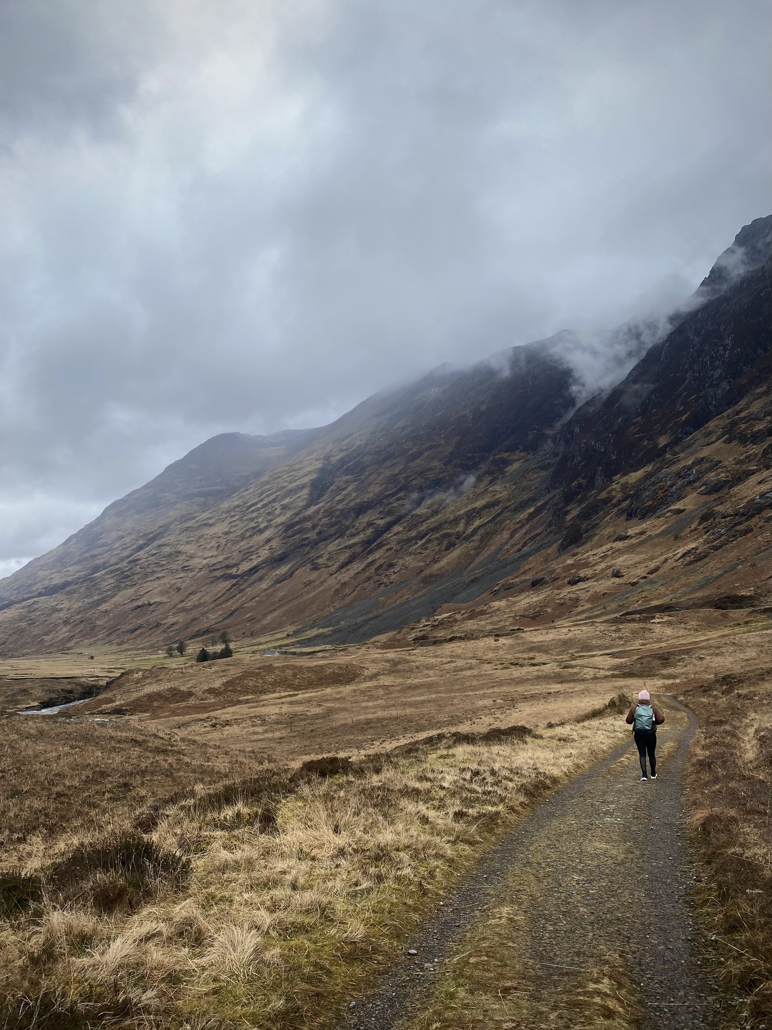 A hiker walking on a dirt trail through a valley with brown grass, with mountains covered in mist and clouds in the background.