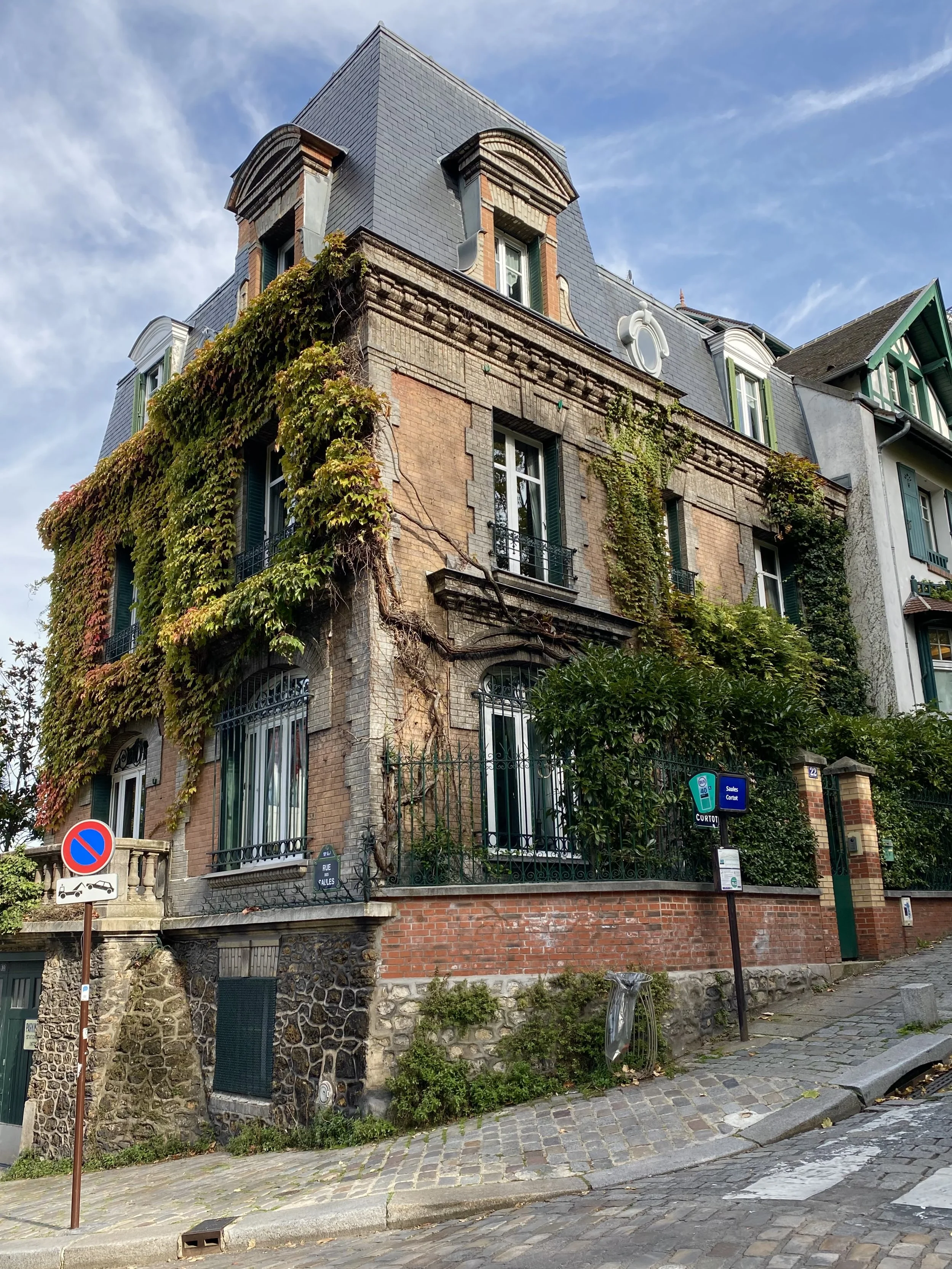 A multi-story building with brick and stone facade, Ivy growing on one side, with French windows and decorative roof dormers, situated on a cobblestone street.