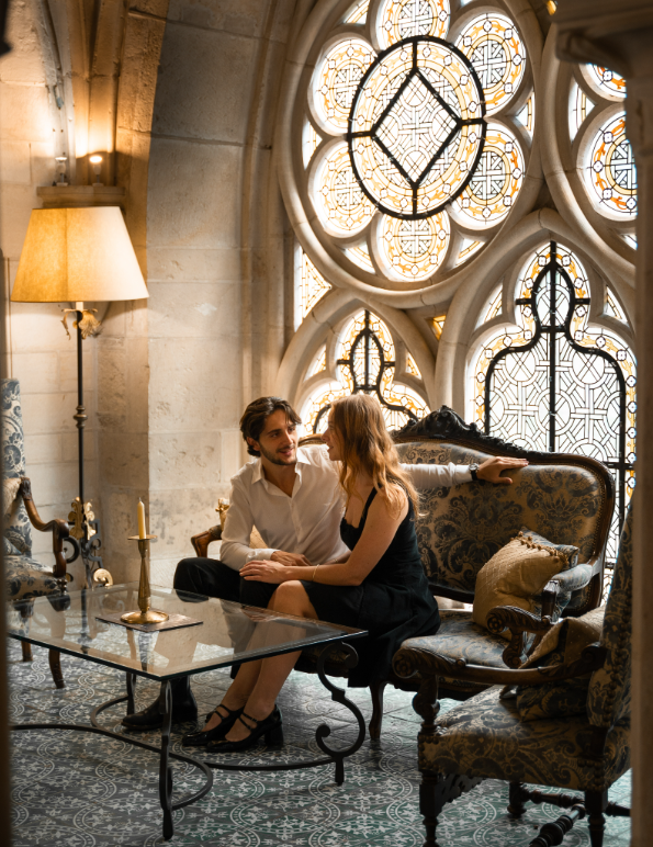 A man and woman sitting on a vintage sofa inside a historic building with stained glass windows and ornate decor, engaged in conversation.