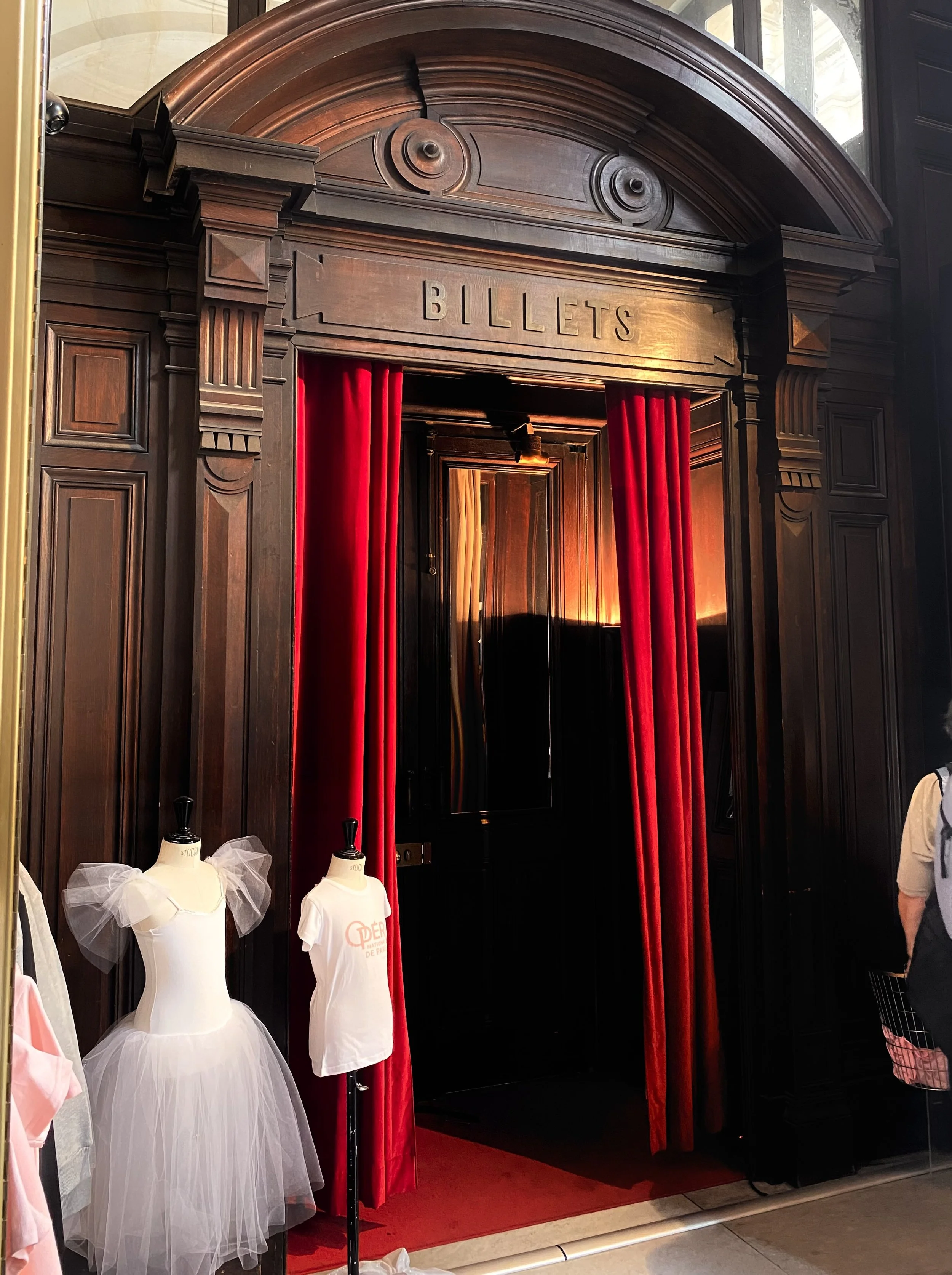 A vintage photo booth with dark wood paneling, red velvet curtains, and mannequins dressed in ballet tutus and t-shirts, set in a classic theater or hotel lobby.