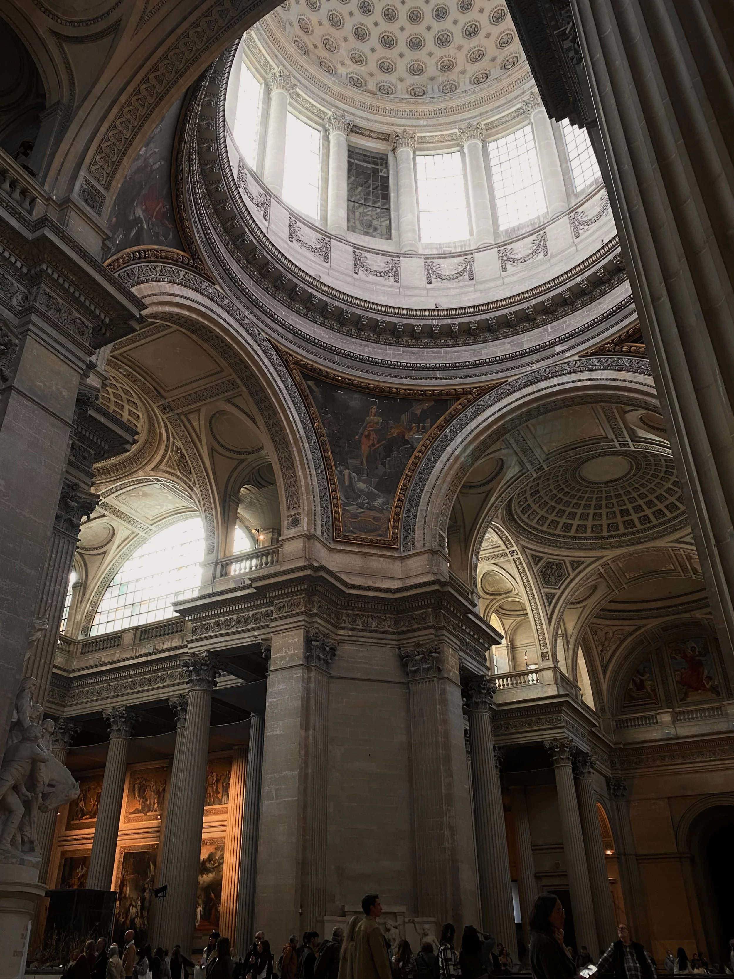 Interior view of a grand historic building with tall columns, arched ceilings, ornate decorations, large windows, and a group of people walking and observing inside.