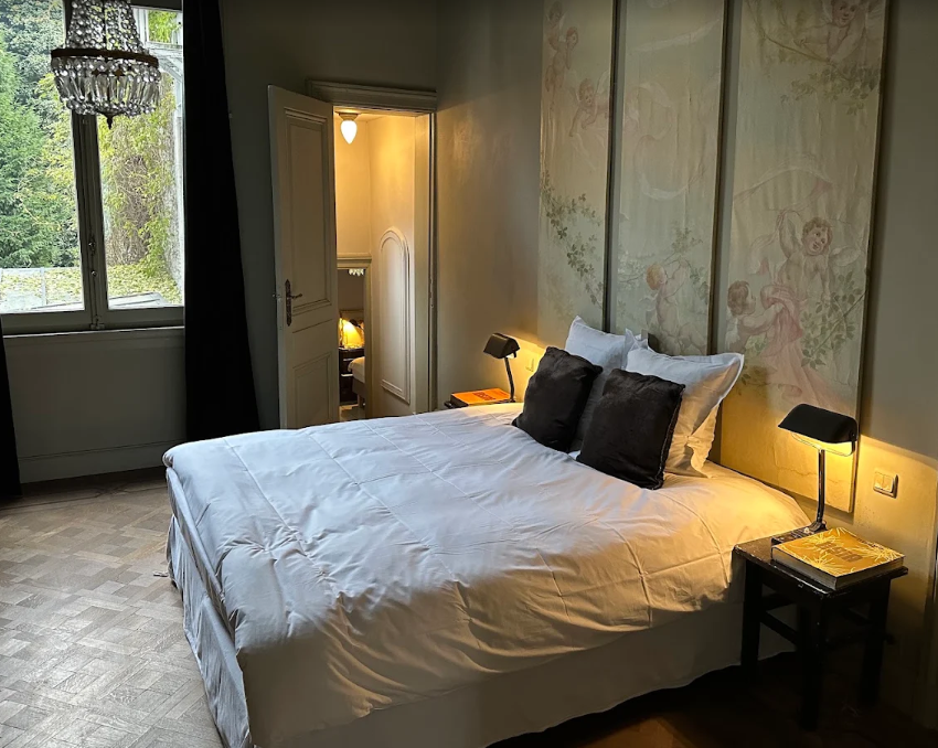 A bedroom with a large bed, white bedding, two black pillows, two white pillows, a black side table with books and a lamp, a window with view of trees, a chandelier, and decorative floral panels on the wall.