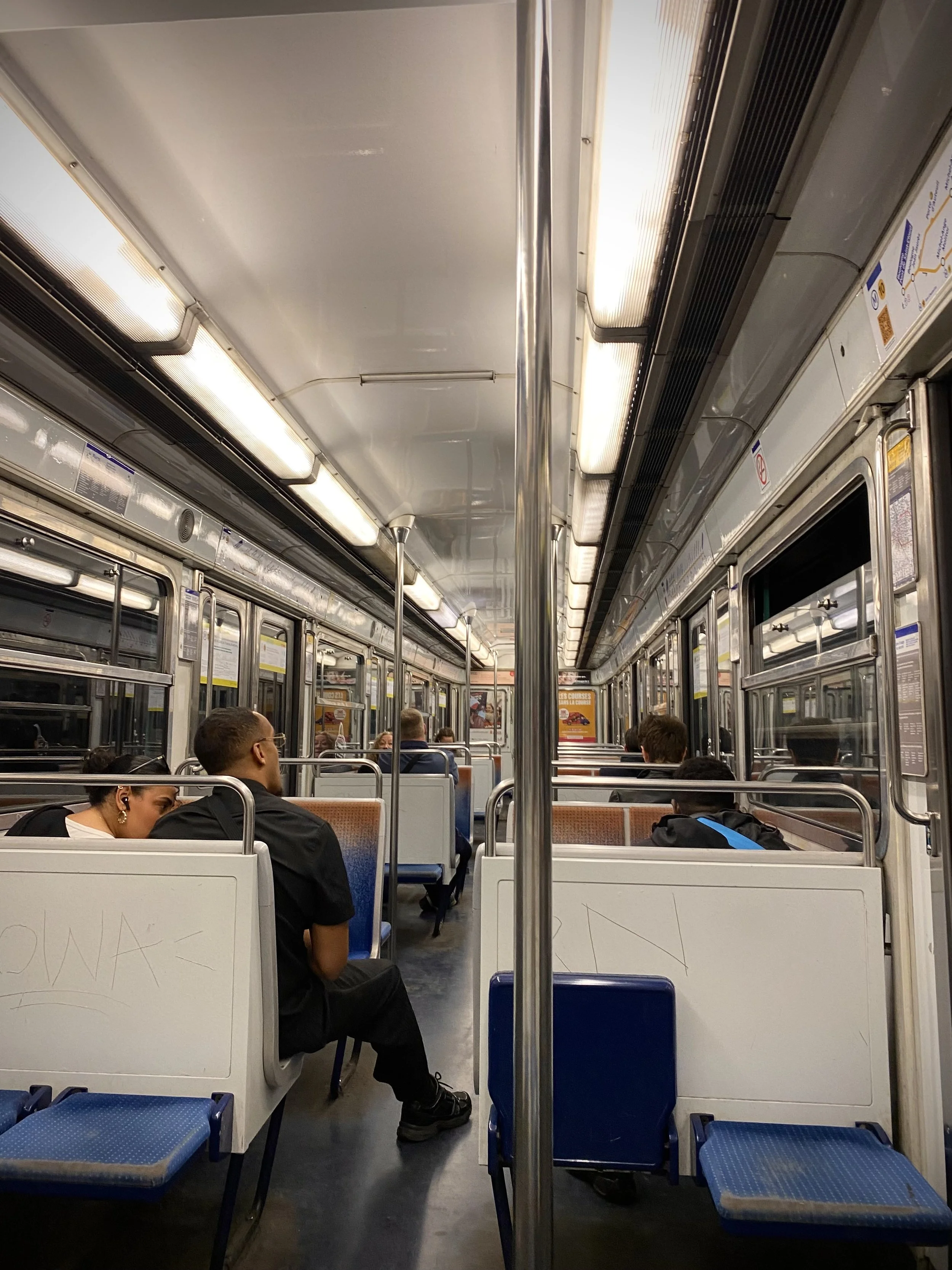 View of the interior of a subway train with passengers seated, overhead lights, metal poles, and windowed doors along the aisle.