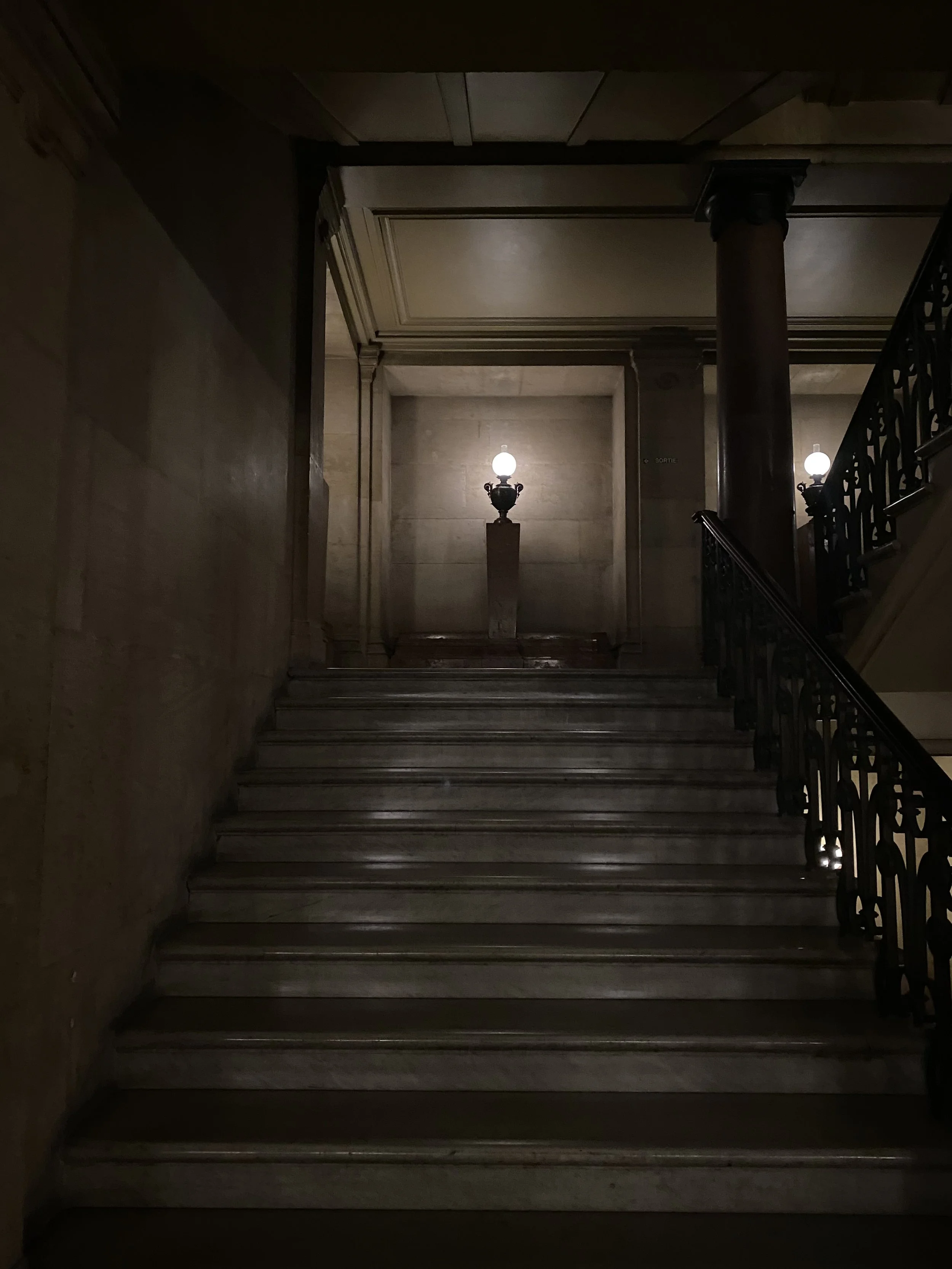 Dimly lit staircase with metal railings on the right, a column in the middle, and three vintage-style bedside lamps on a ledge at the top, illuminating the wall.