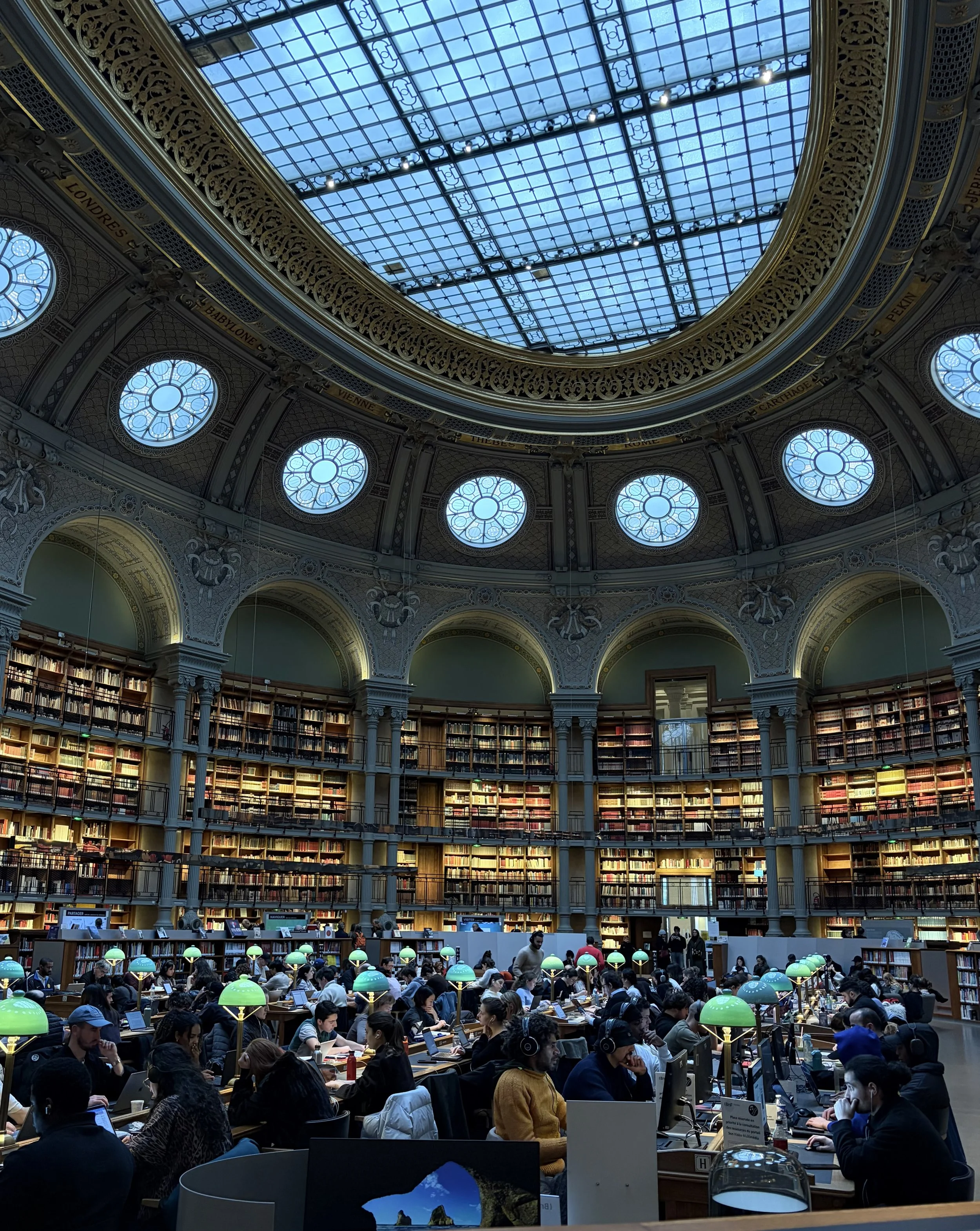 View of a large, multi-level library with a domed glass ceiling, filled with people sitting at long tables working on laptops and reading. The upper shelves are lined with books, and the scene is lively with students and visitors.