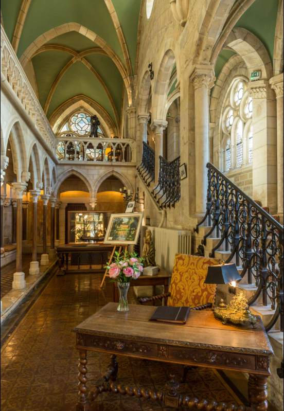Interior of a historic building with stone arches, stained glass windows, and wooden furniture, including a desk with a lamp and a bouquet of pink roses, staircase with ornate iron railing, and artworks displayed on easels.