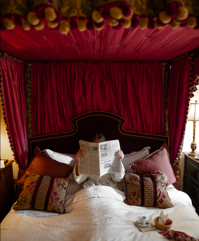 Person in bed reading a newspaper in a luxurious bedroom with red drapes and ornate pillows.