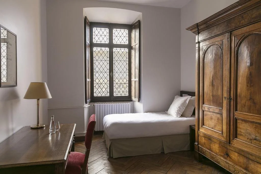 A small bedroom with a single bed, a wooden wardrobe, a desk with a lamp, and a window with open wooden shutters.