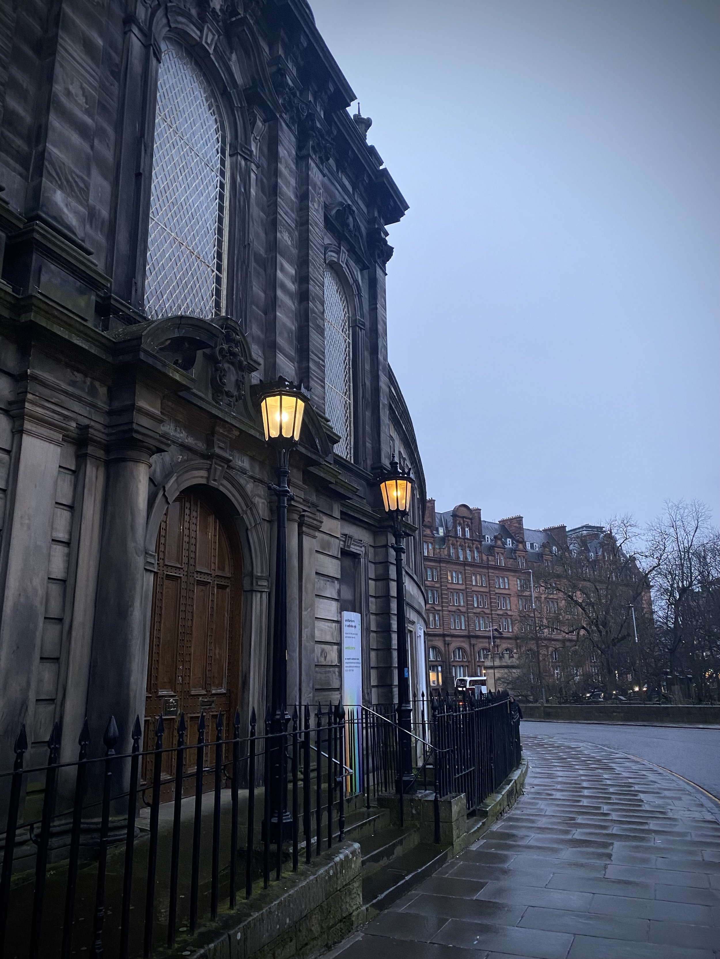 A historic stone building with large decorative windows and wooden doors, illuminated by two street lamps, on a rainy evening in an urban area with other brick buildings in the background and leafless trees.