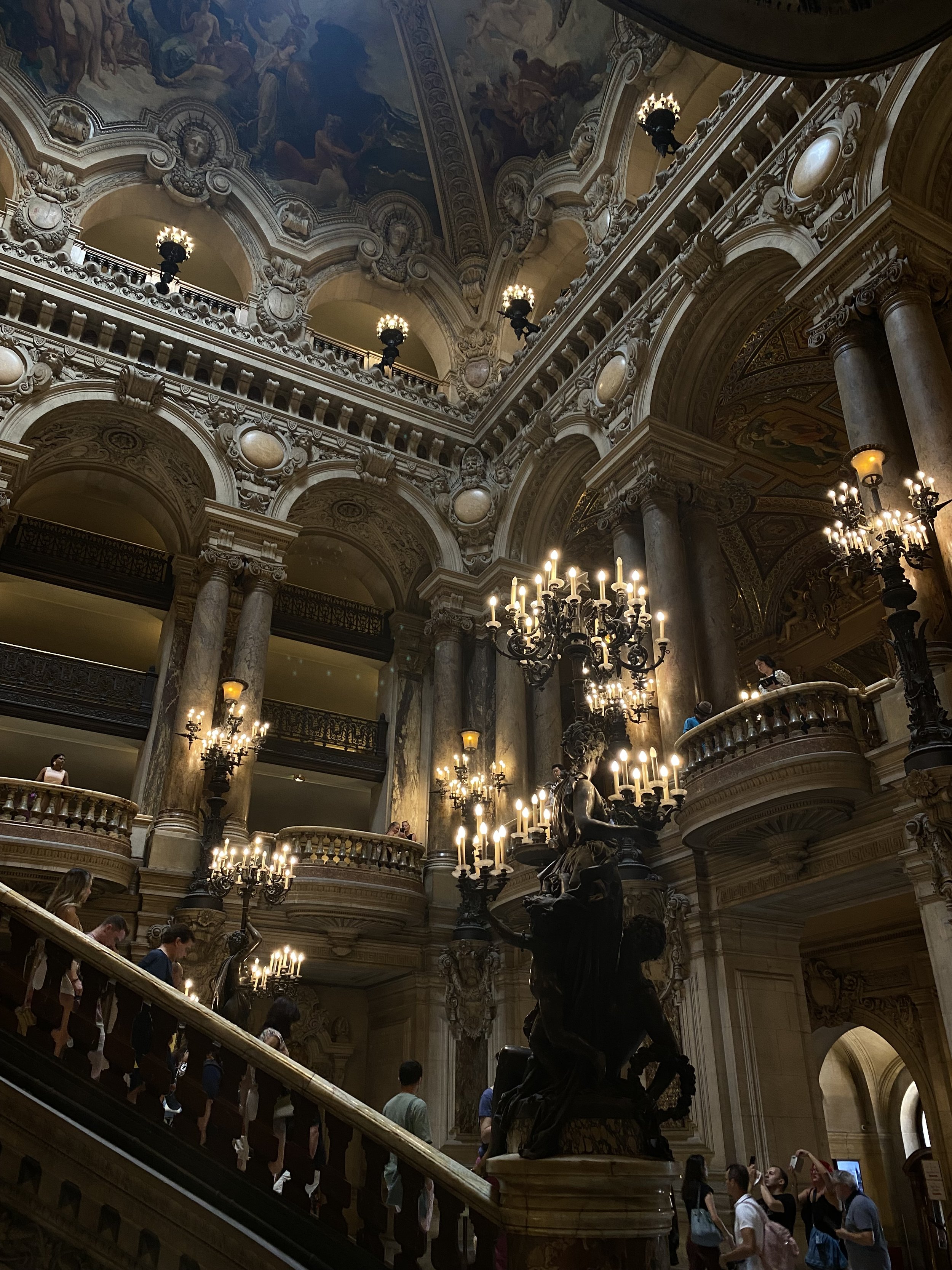 Interior of a grand historic building with ornate architecture, gold accents, chandeliers, and visitors walking on the staircase and inside the building.