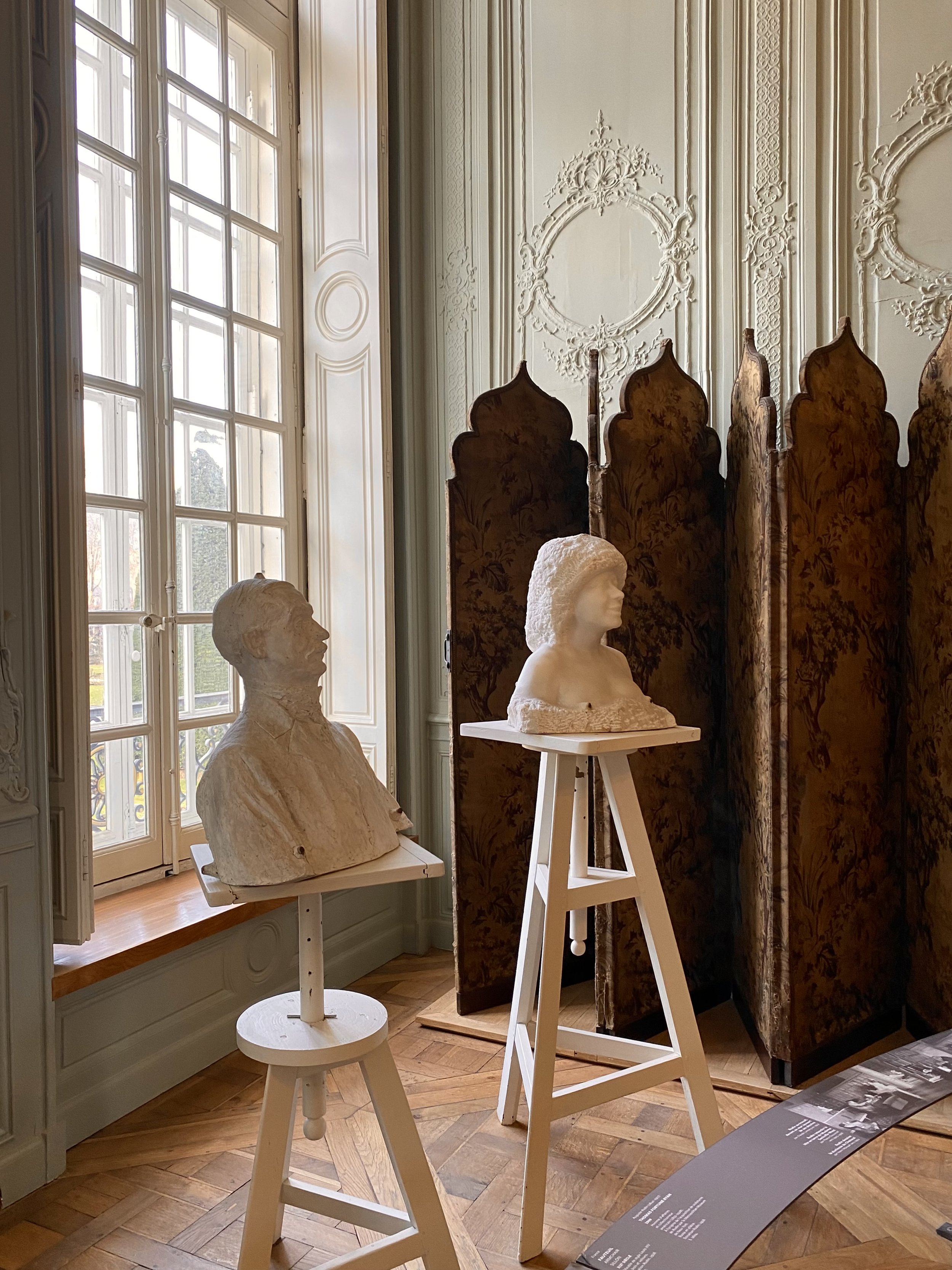 Art exhibit with bust sculptures on stools and a folding screen, located near a large window with daylight.