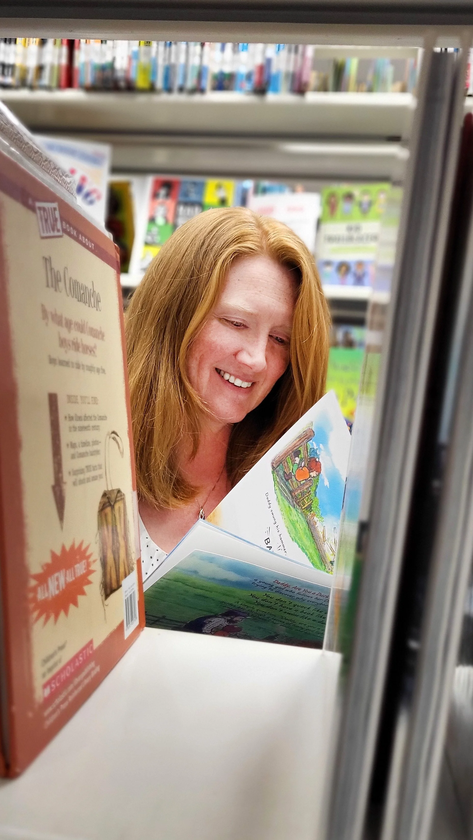 A woman with red hair reading a children's book on a store shelf, surrounded by other books.