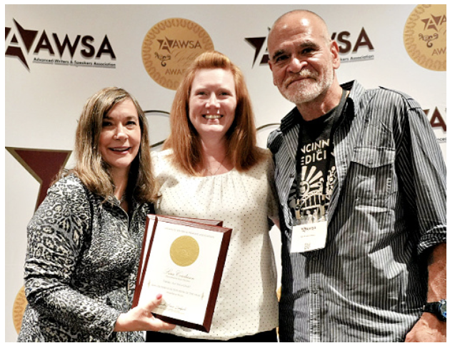 Three people standing together at an awards event, with two women and one man, smiling, and one woman holding a framed certificate. The background features banners with the text 'AWSA' and a logo.