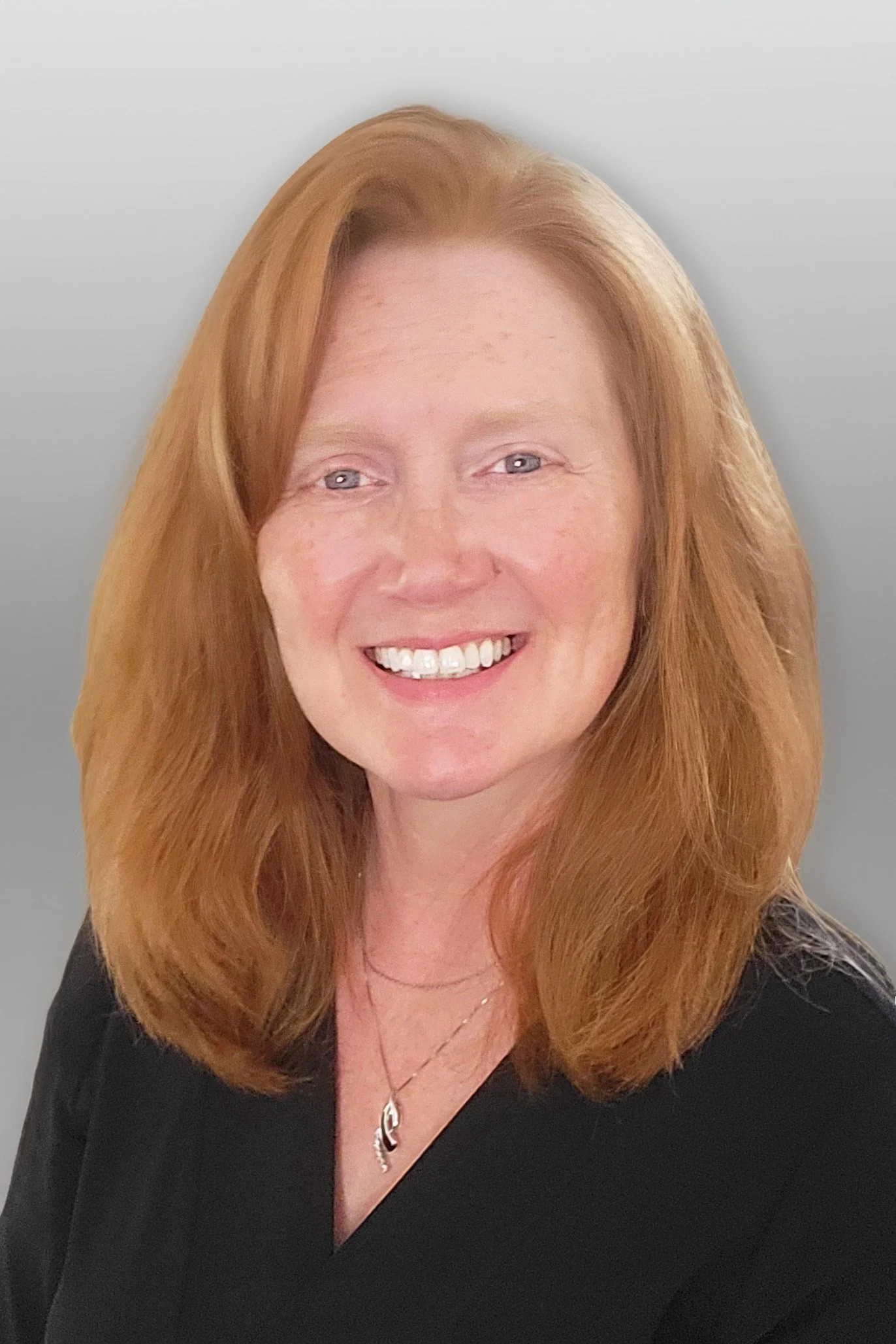Portrait of a smiling woman with red hair, wearing a black top and a silver necklace, against a light gray background.