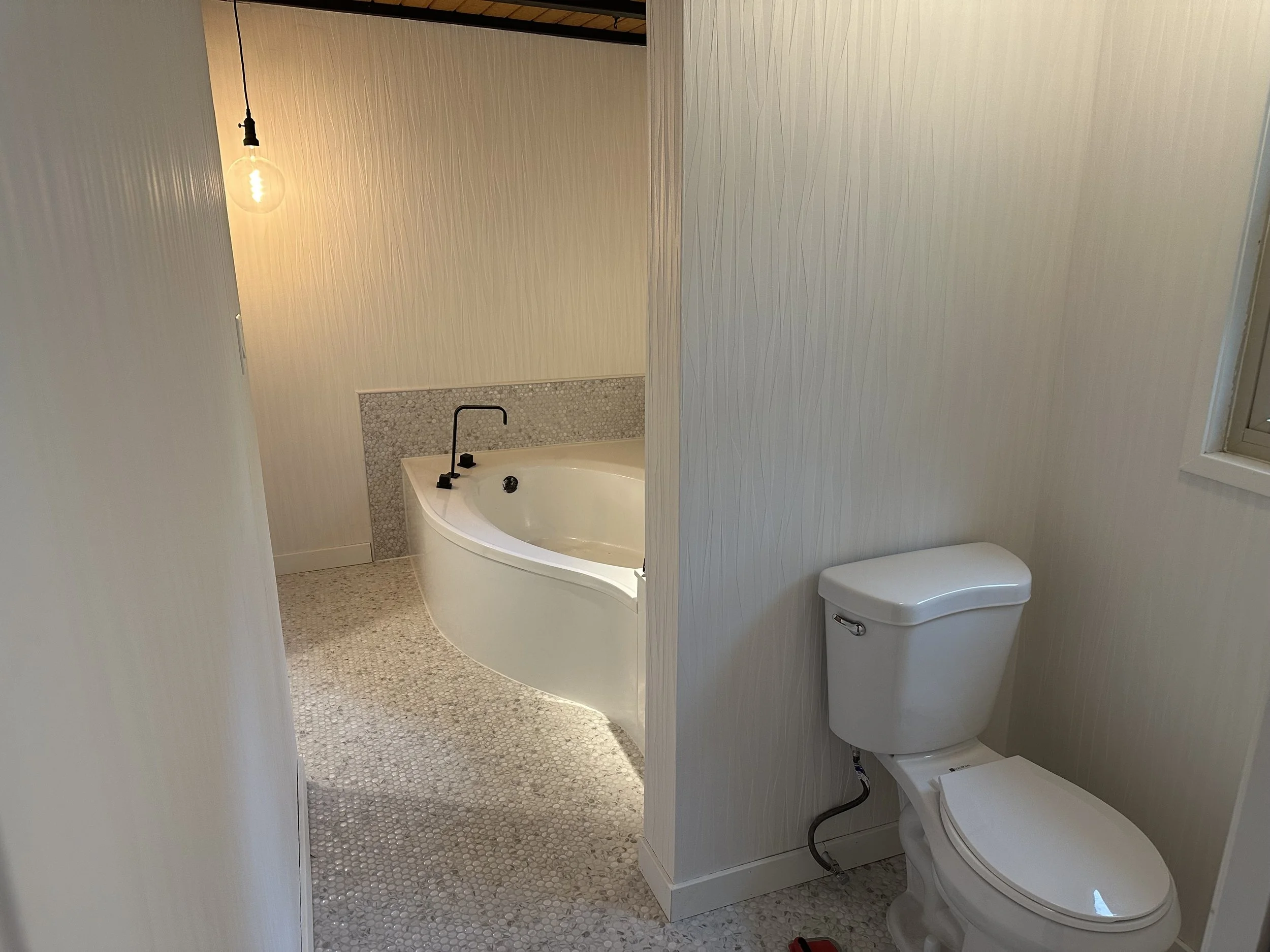 Bathroom with a corner bathtub, a wall with textured wallpaper, a hanging lightbulb, and a toilet.