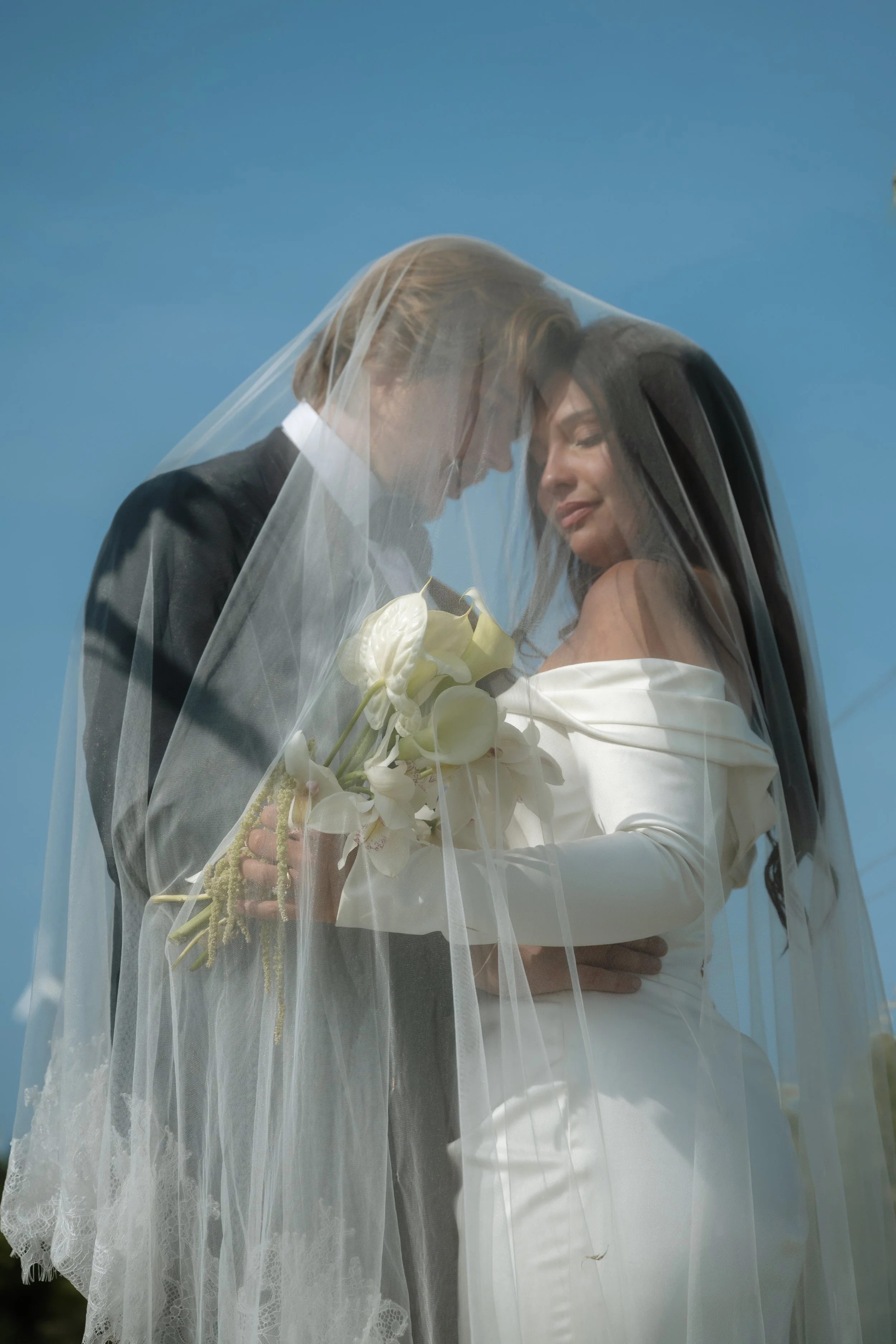 A bride and groom under a wedding veil, holding a bouquet of white flowers, with a blue sky in the background.