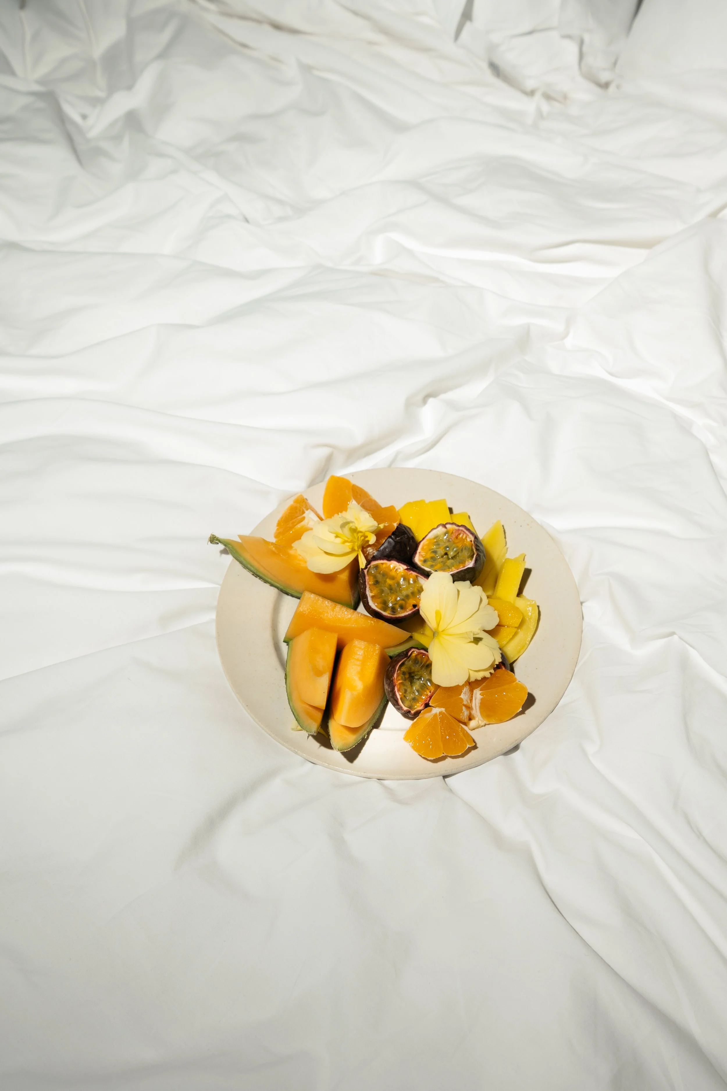 A plate of assorted fresh tropical fruits including watermelon, cantaloupe, orange slices, and passion fruit, garnished with edible flowers, set on a white surface.