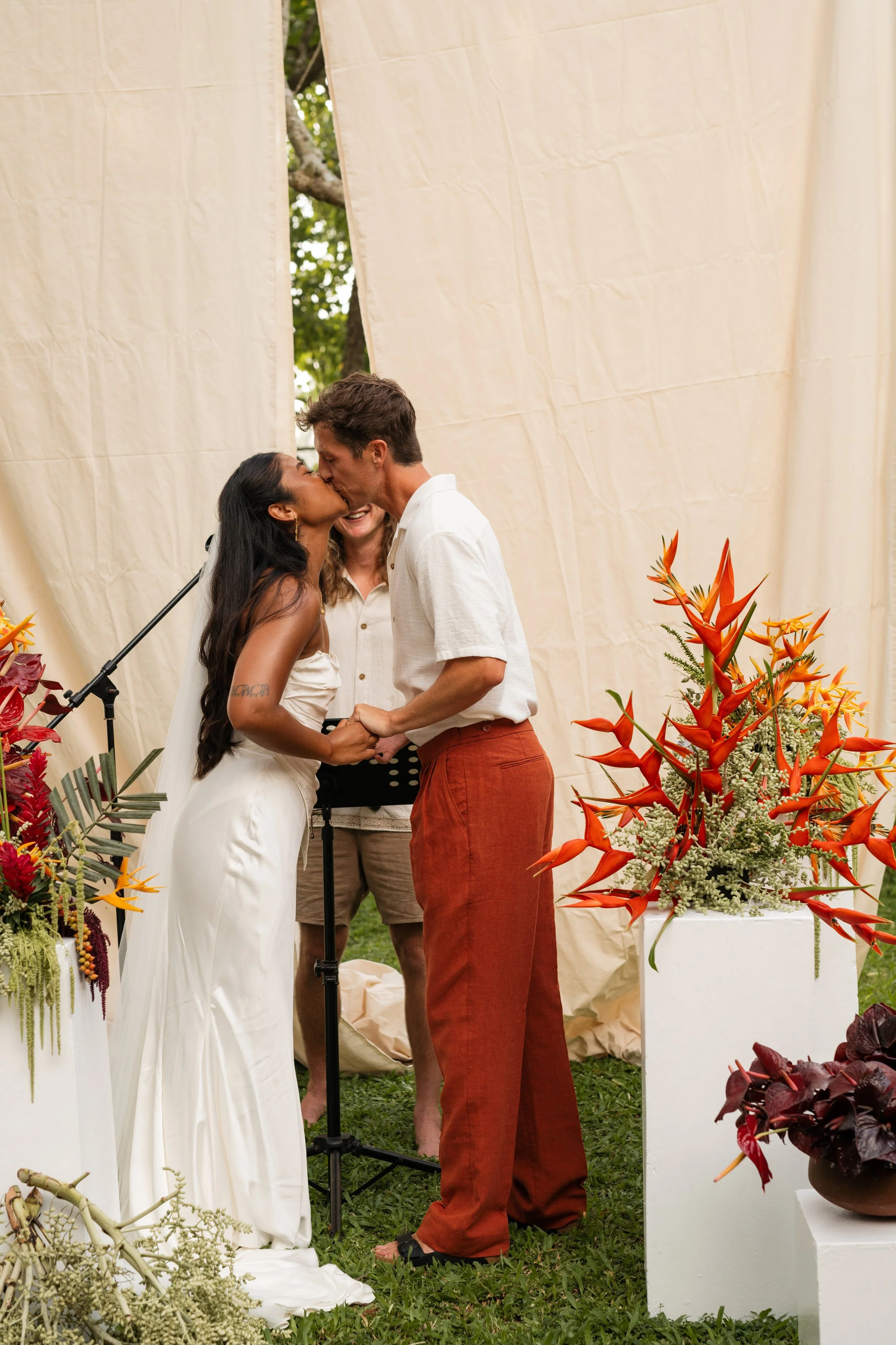 A couple kissing during their wedding ceremony, with a woman officiating, surrounded by floral decorations and a cream-colored backdrop outdoors.