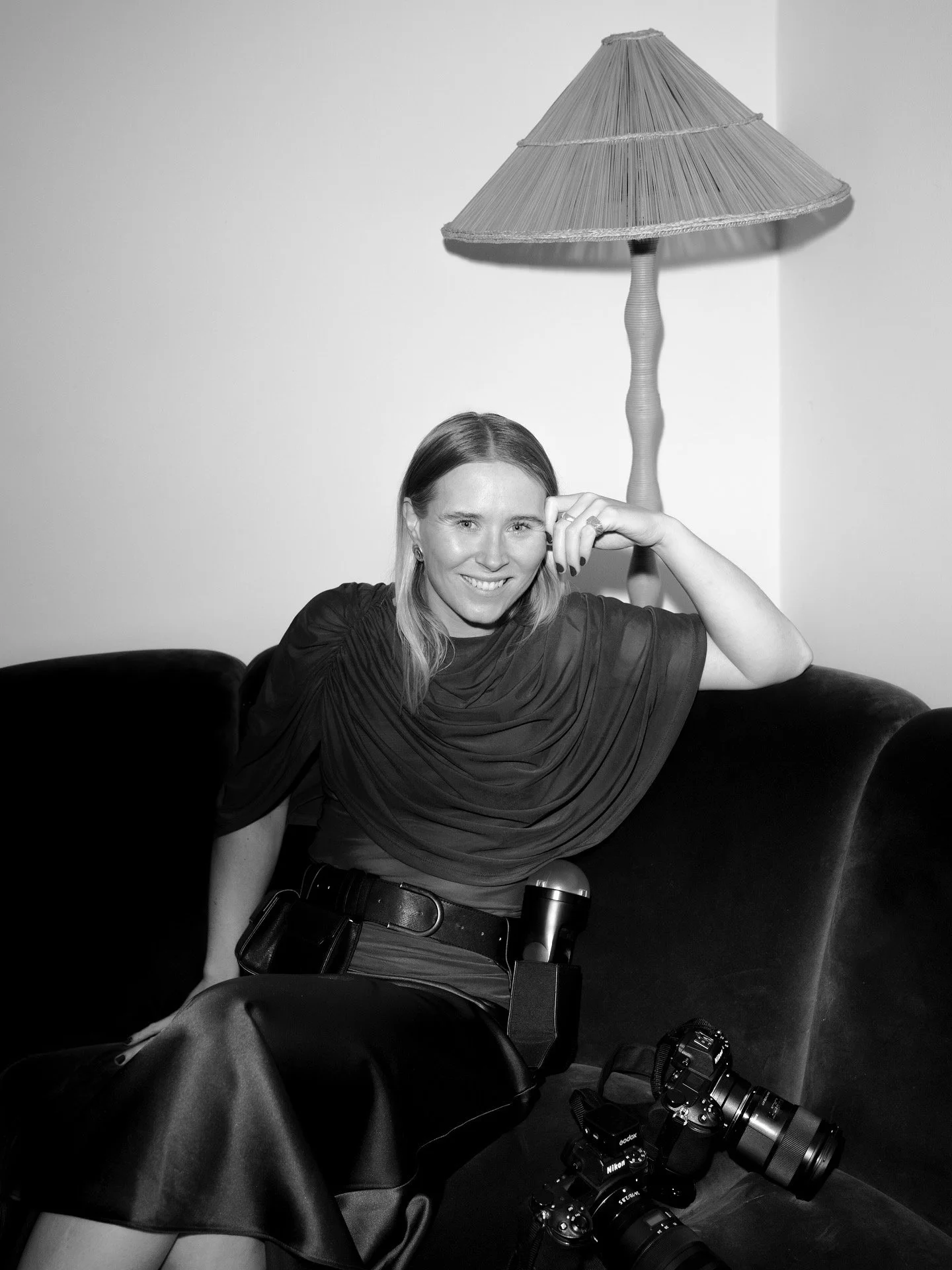 Black and white photo of a woman sitting on a sofa, smiling at the camera. She has a camera around her neck, a microphone in her lap, and several cameras on the sofa next to her. A large lampshade hangs behind her.