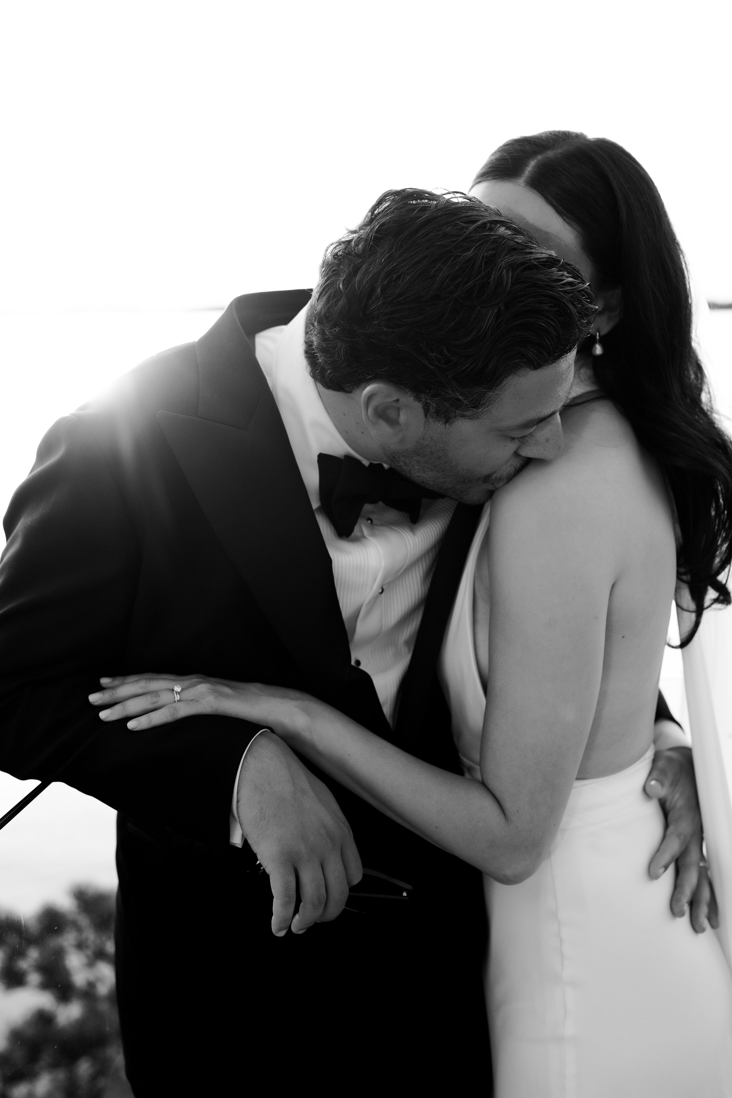 A black and white photo of a bride and groom sharing a wedding kiss, with the groom in a tuxedo and the bride in a sleeveless dress, outdoors with a blurred natural background.