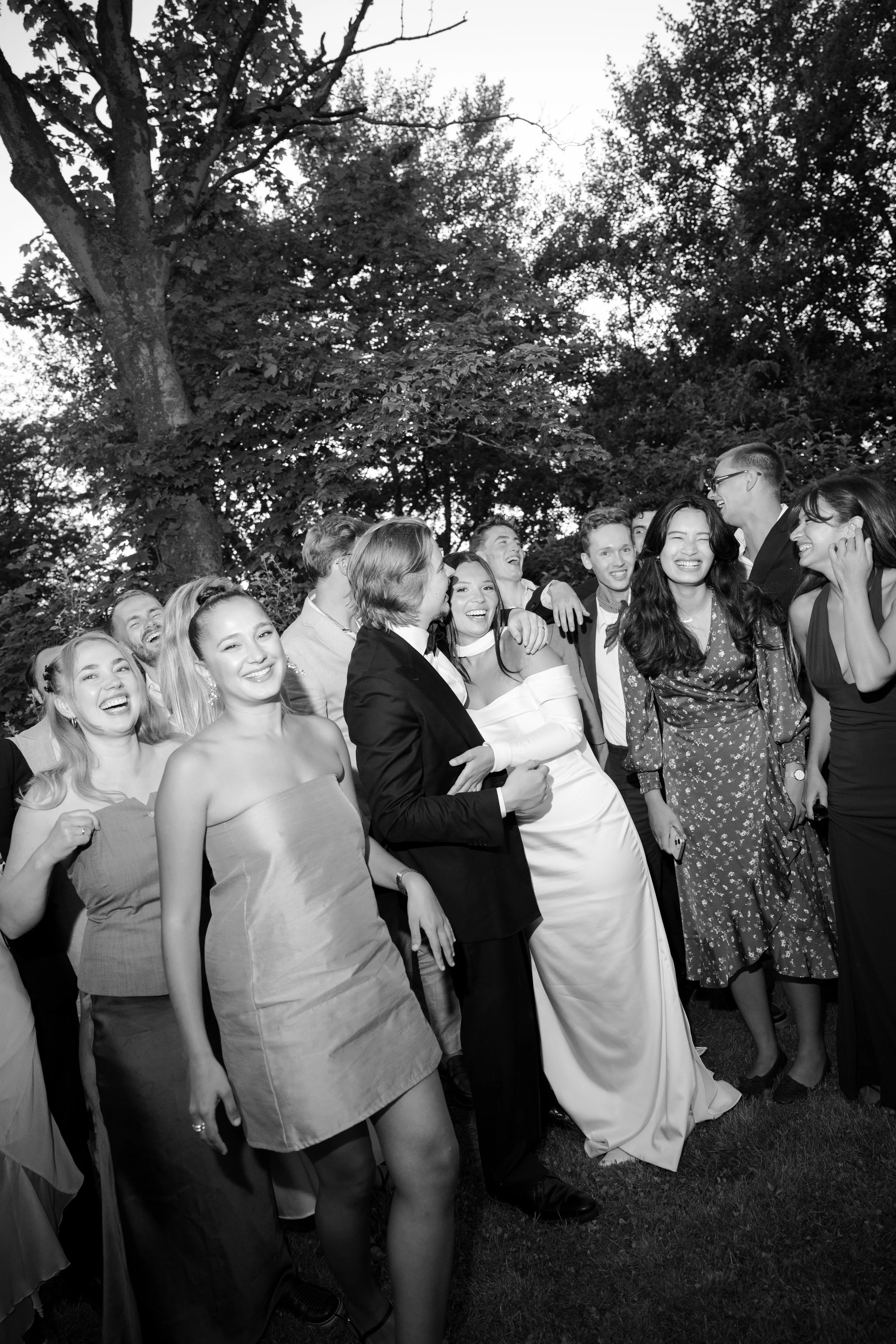 Group of people dressed in formal attire laughing and enjoying outdoors under trees.