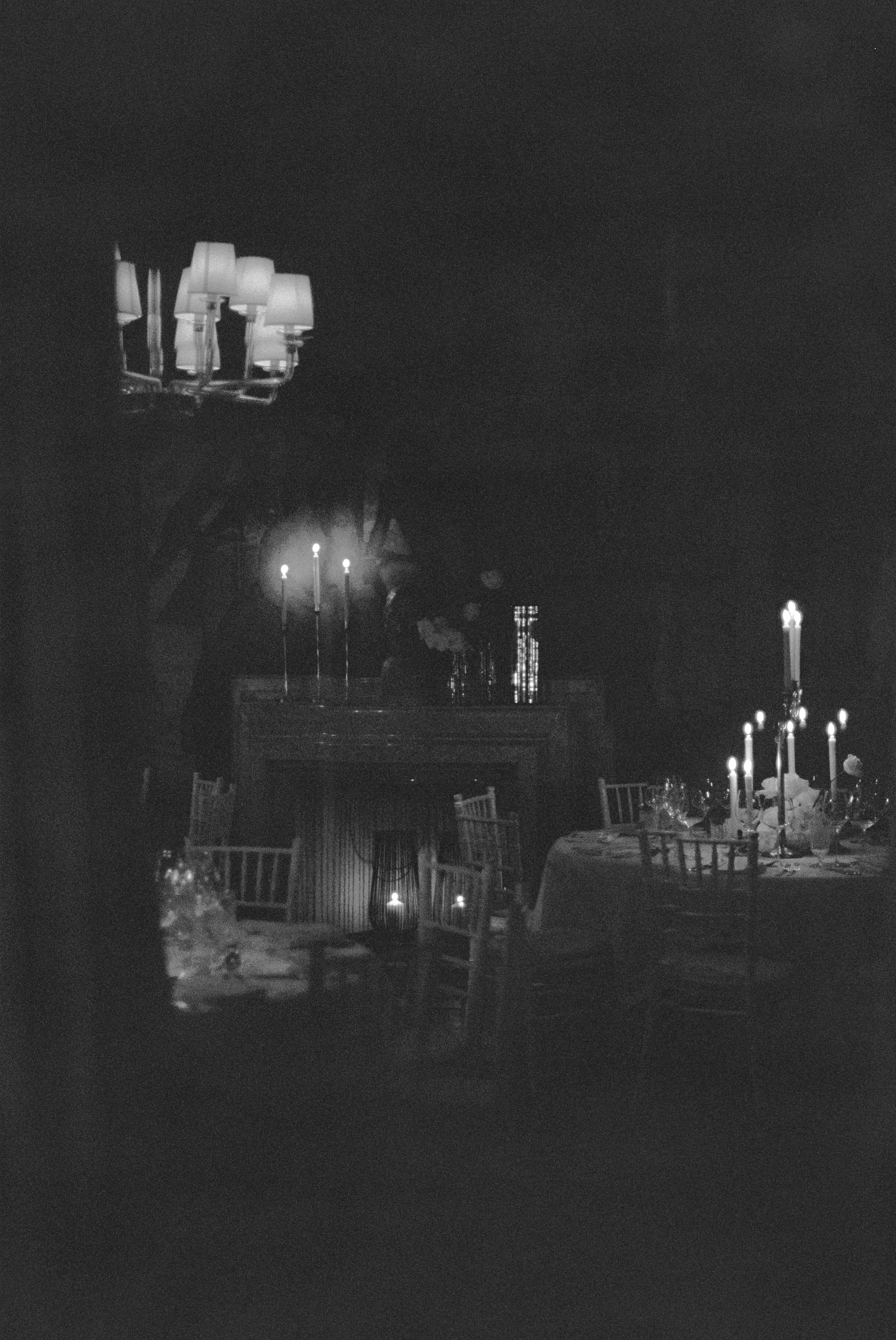 A dimly lit dining room with candles on the table and a mantelpiece in the background. A chandelier with lamps is visible at the top.