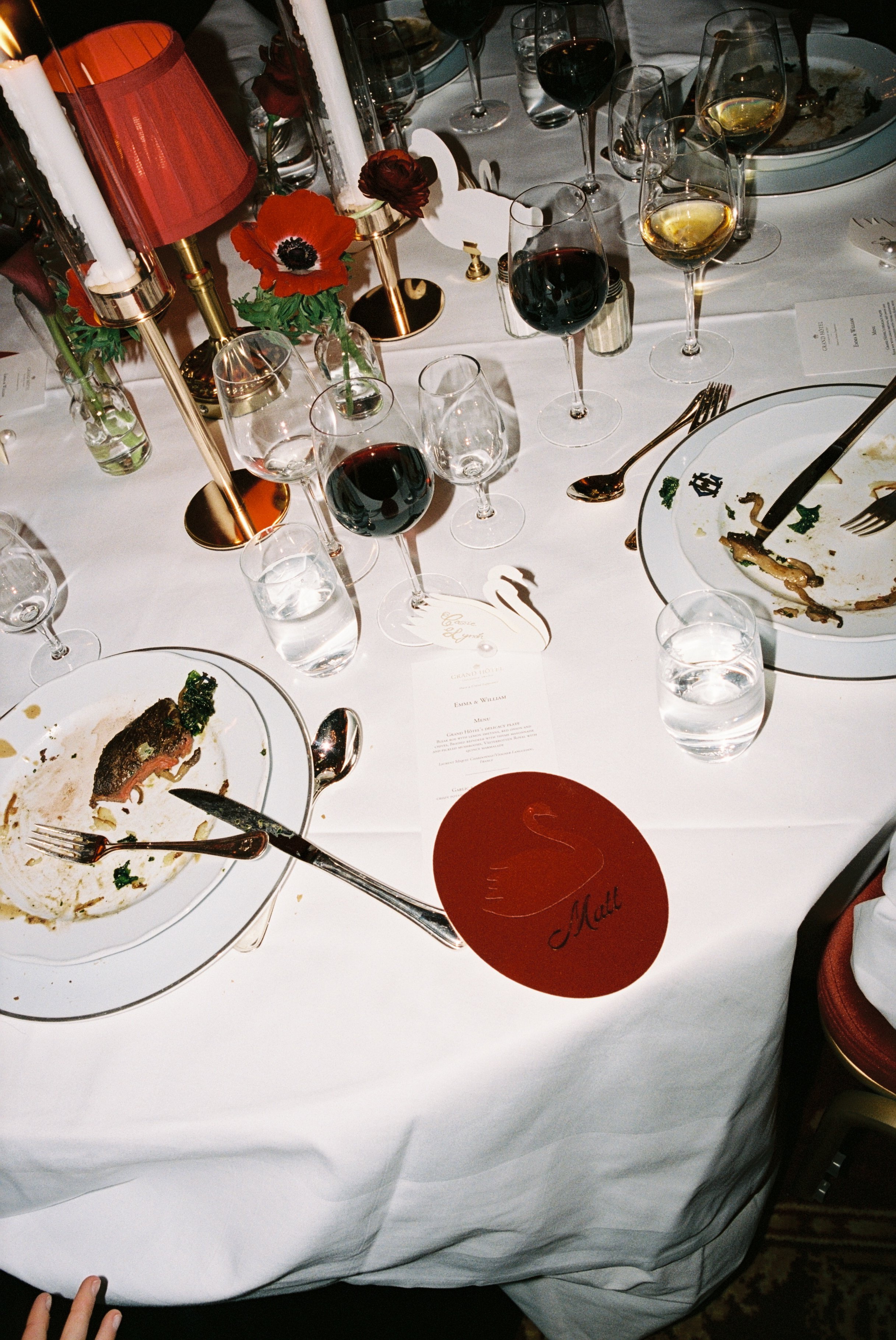 A dining table with empty plates, glasses of red and white wine, water glasses, candles, and red flowers in vases, indicating a finished meal at a formal event.