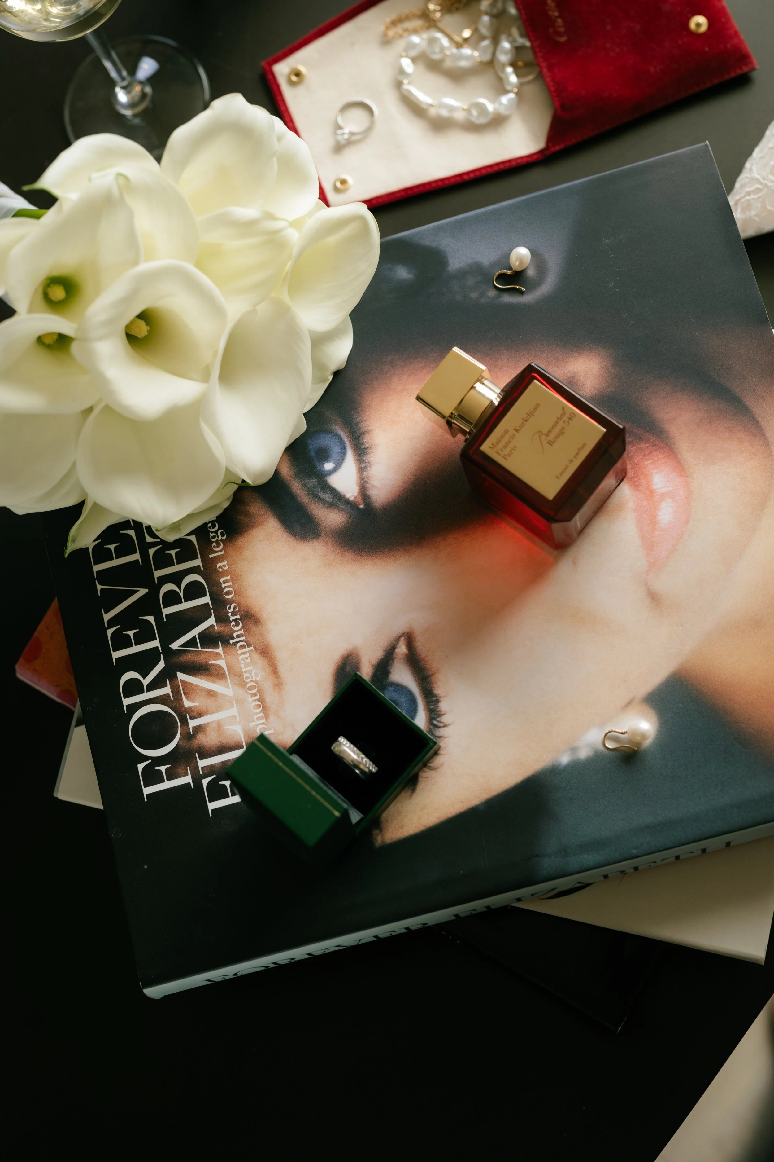A bouquet of white calla lilies on top of a fashion magazine featuring a woman's face, with jewelry, perfume, and earrings arranged around it.